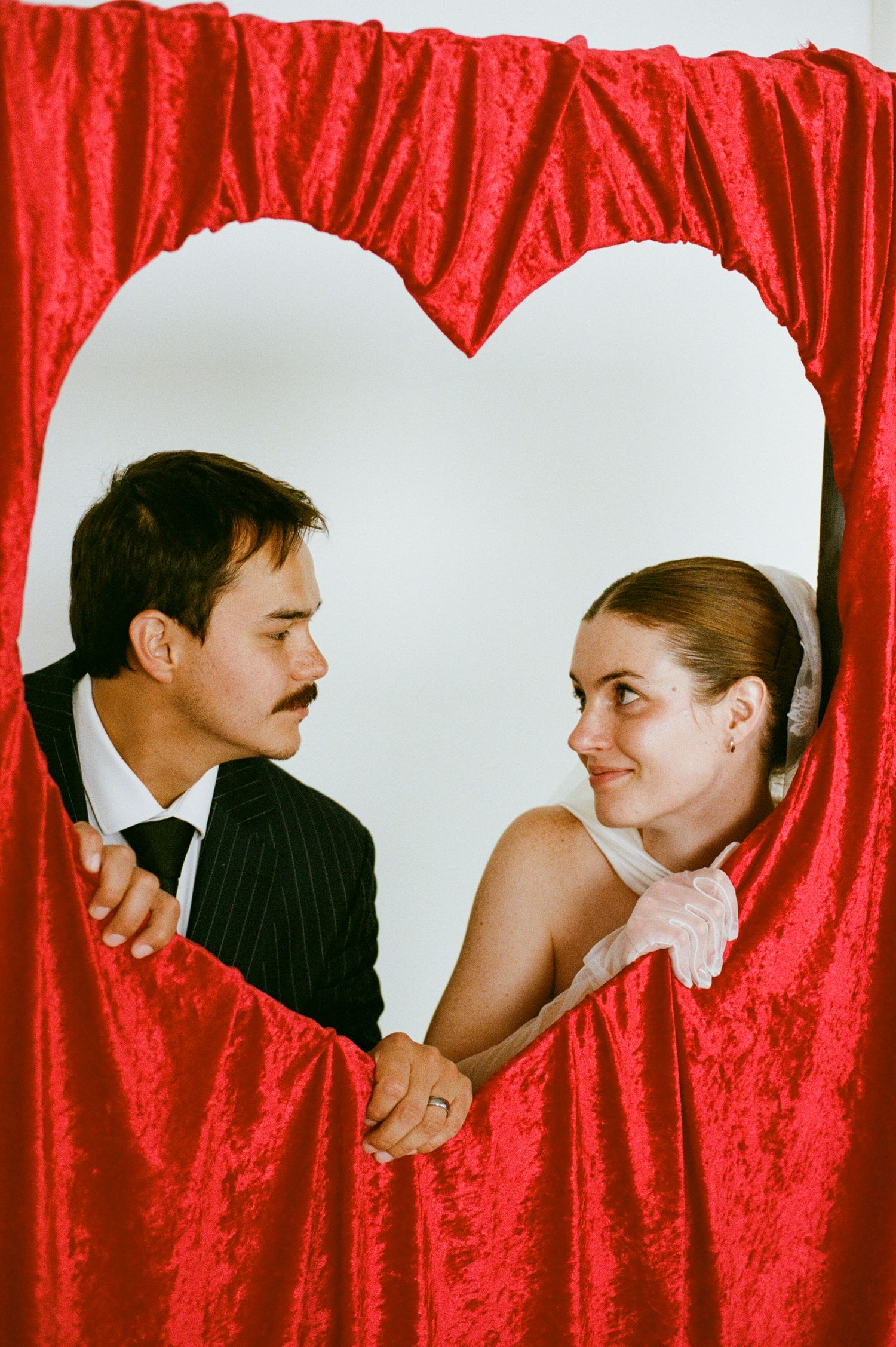 Couple holding hands and gazing at each other through a heart-shaped opening in red velvet curtains.