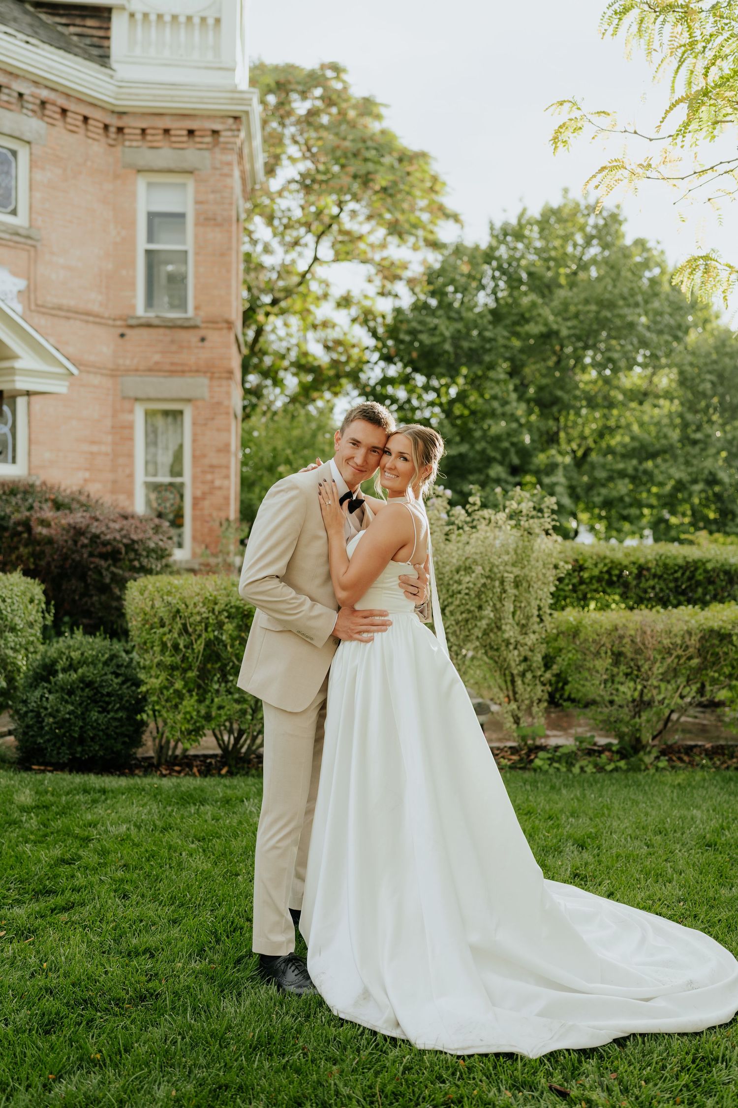 Bride in white gown with train and groom in tan suit embracing outside historic mansion.