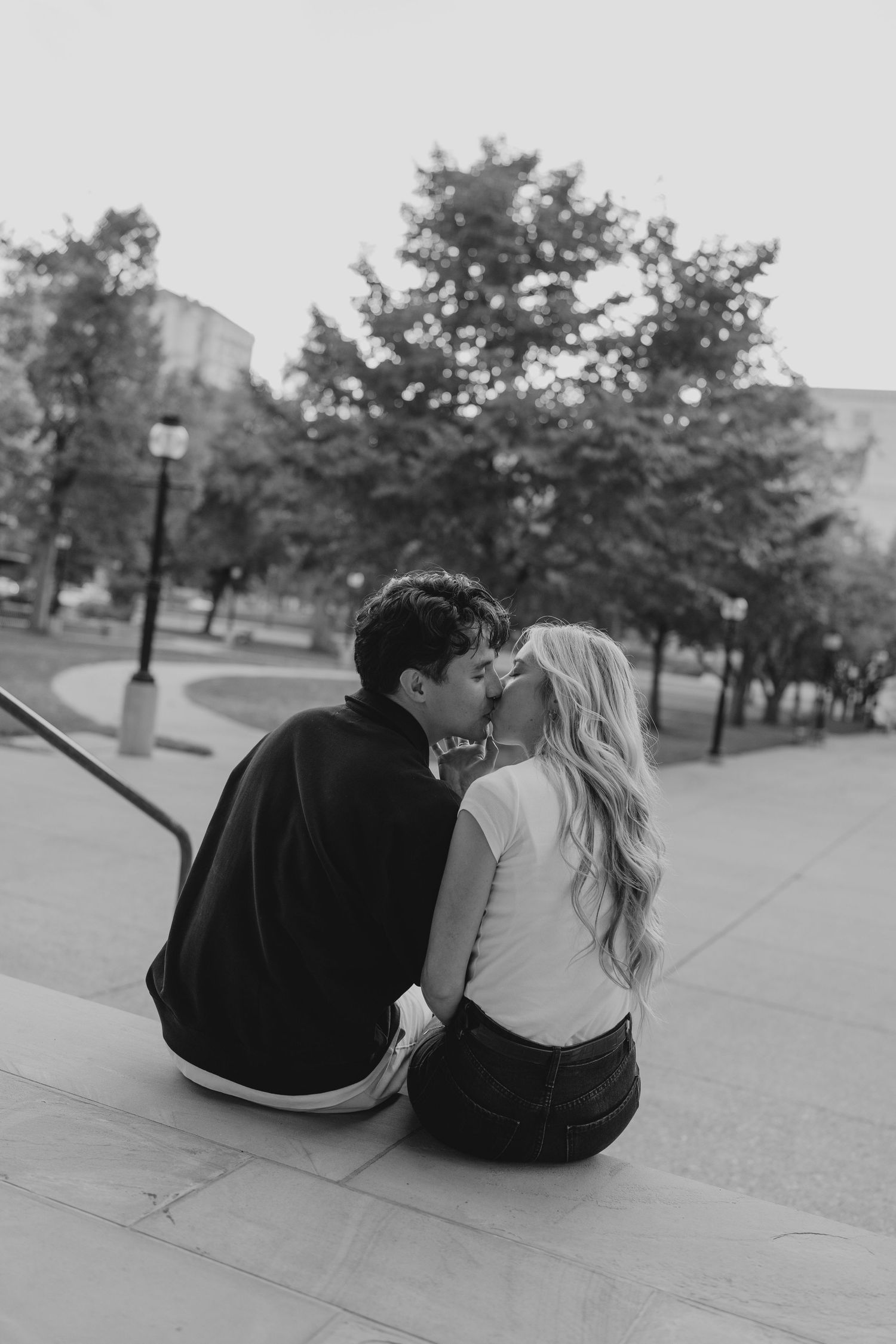 Black and white photo of couple sharing romantic kiss while sitting on outdoor steps.