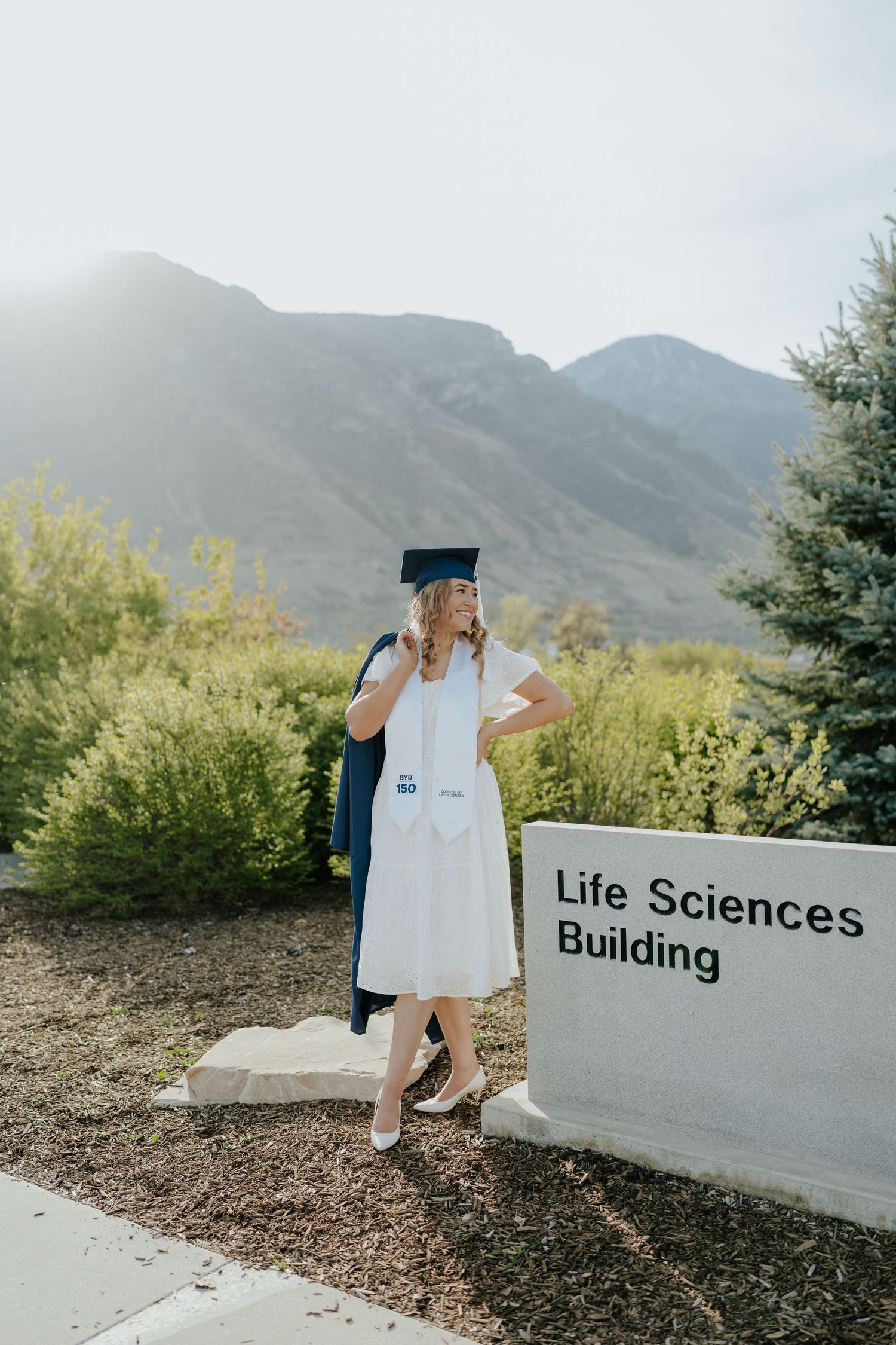 Graduate in white dress and cap poses by Life Sciences Building sign with mountain backdrop.