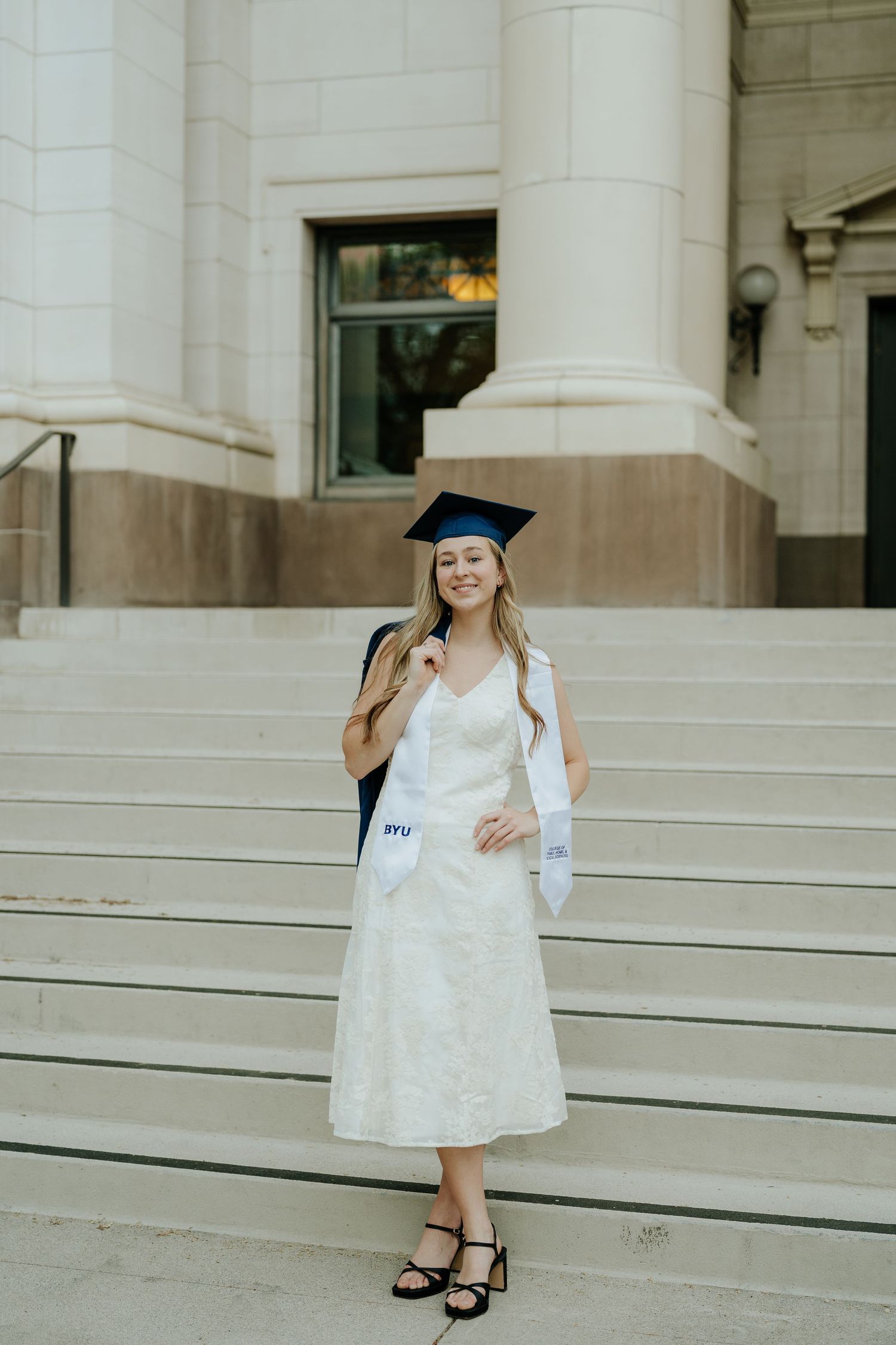 Smiling BYU graduate in white lace dress and cap on stone steps holding graduation regalia.