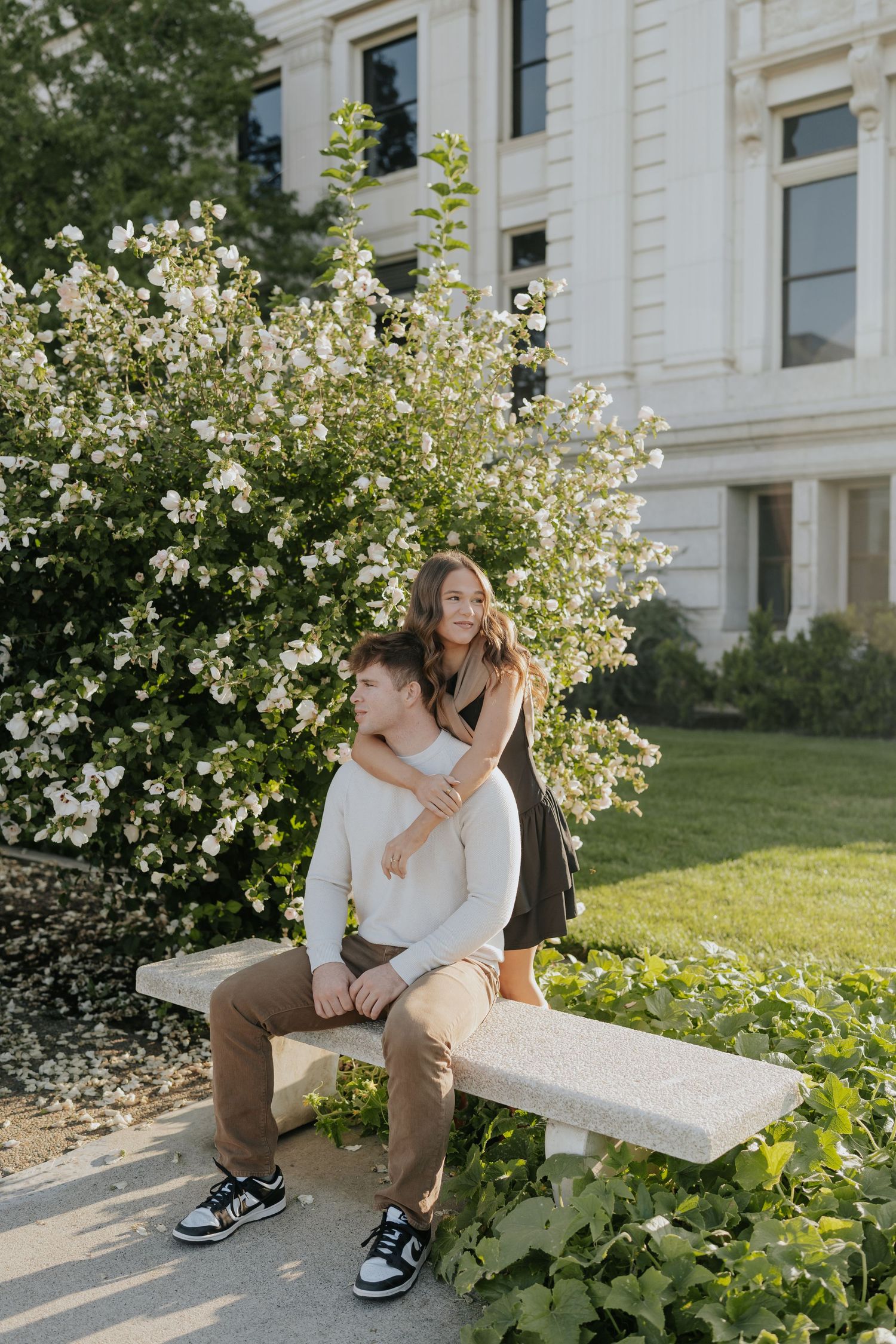 Couple poses near flowering bush, man sitting on stone bench while woman stands behind him embracing.