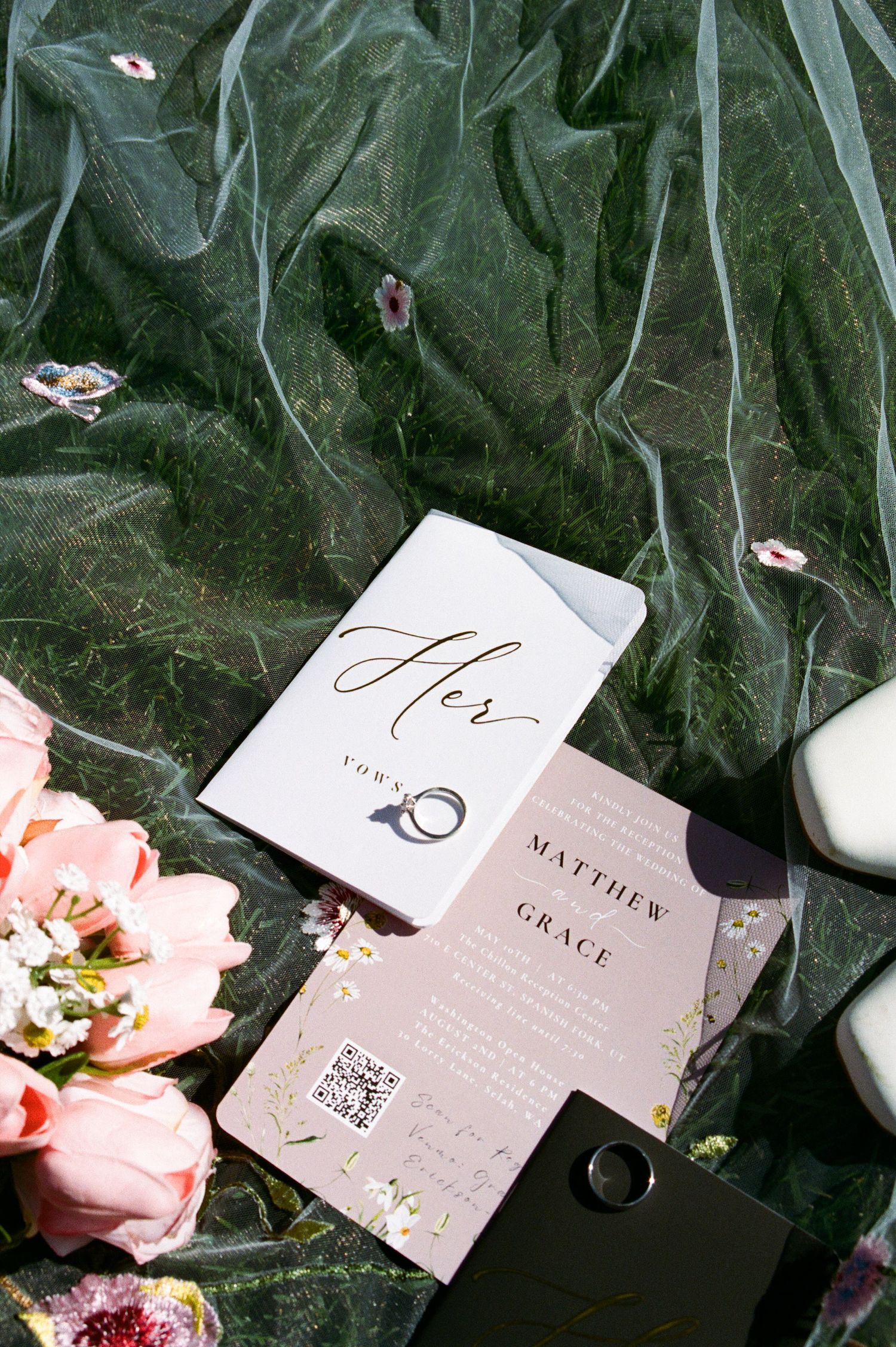 Wedding stationery suite for Matthew and Grace displayed on green tulle with pink flowers and a ring.