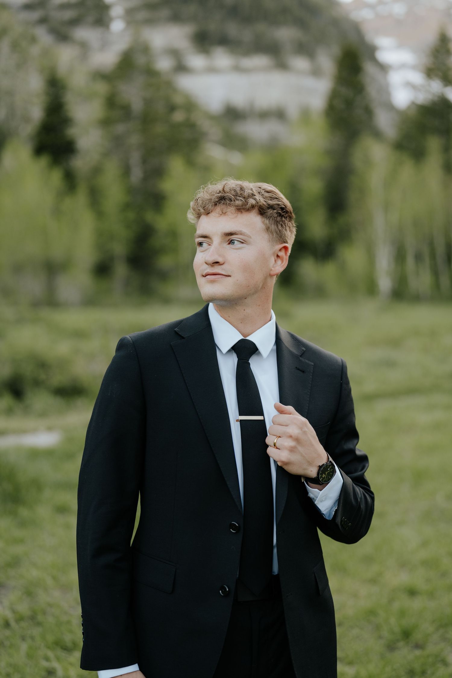 Young man in black suit and tie standing outdoors with green trees and mountains in the background.
