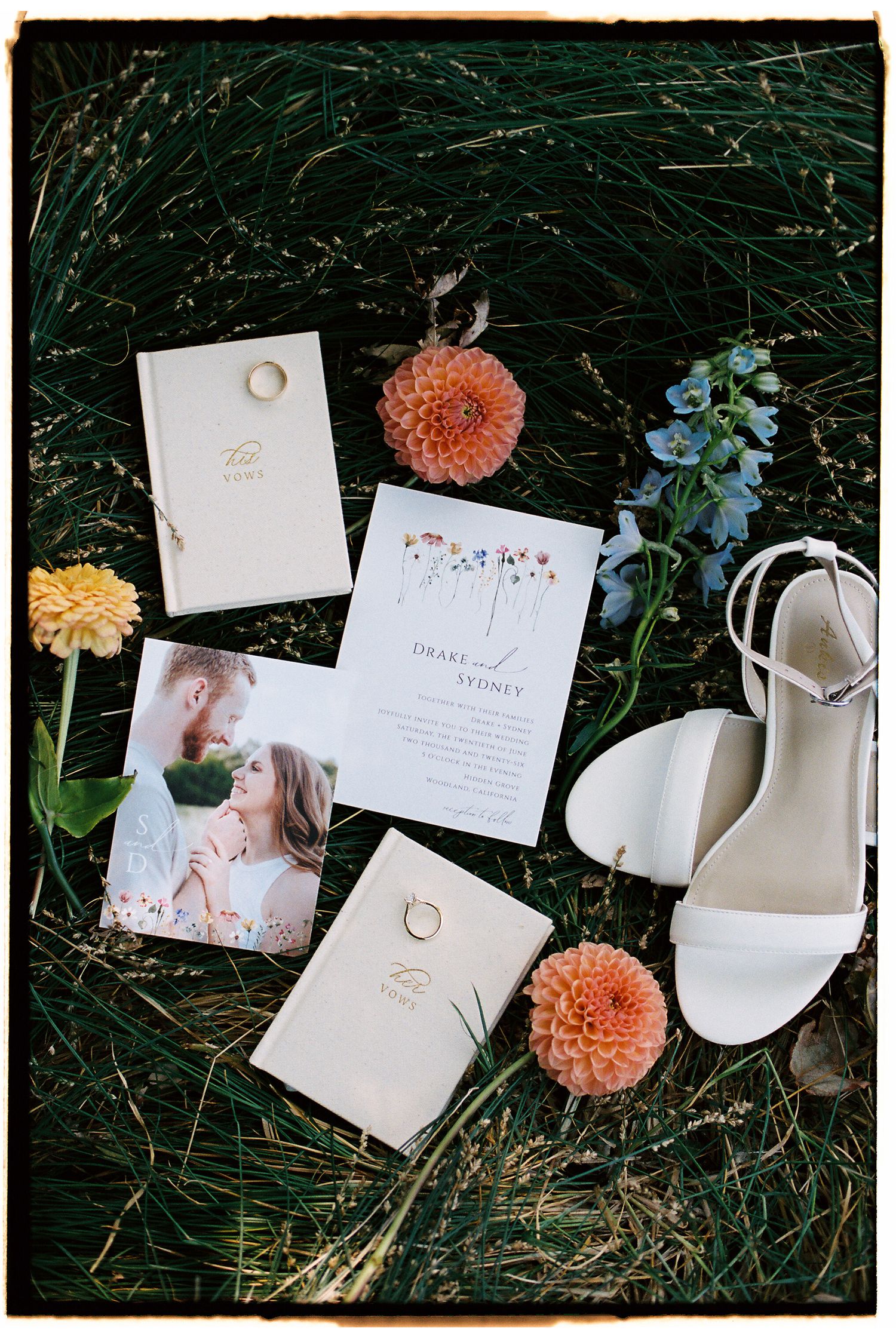 Wedding flat lay on grass featuring invitation suite, gold rings, white heels, orange dahlias, and a romantic couple photo.