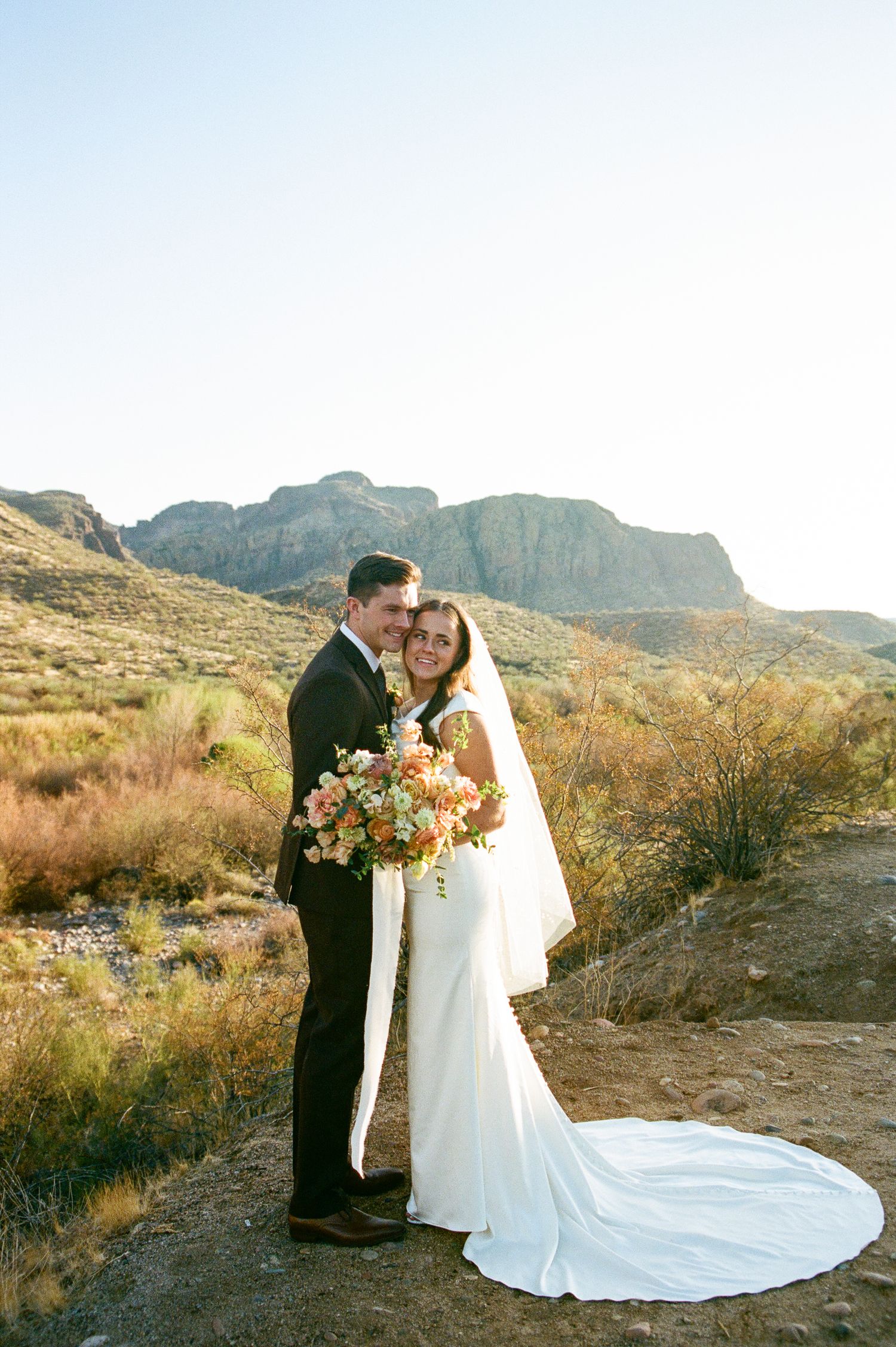 Bride and groom embrace in a desert landscape with dramatic mountain backdrop and colorful bridal bouquet.