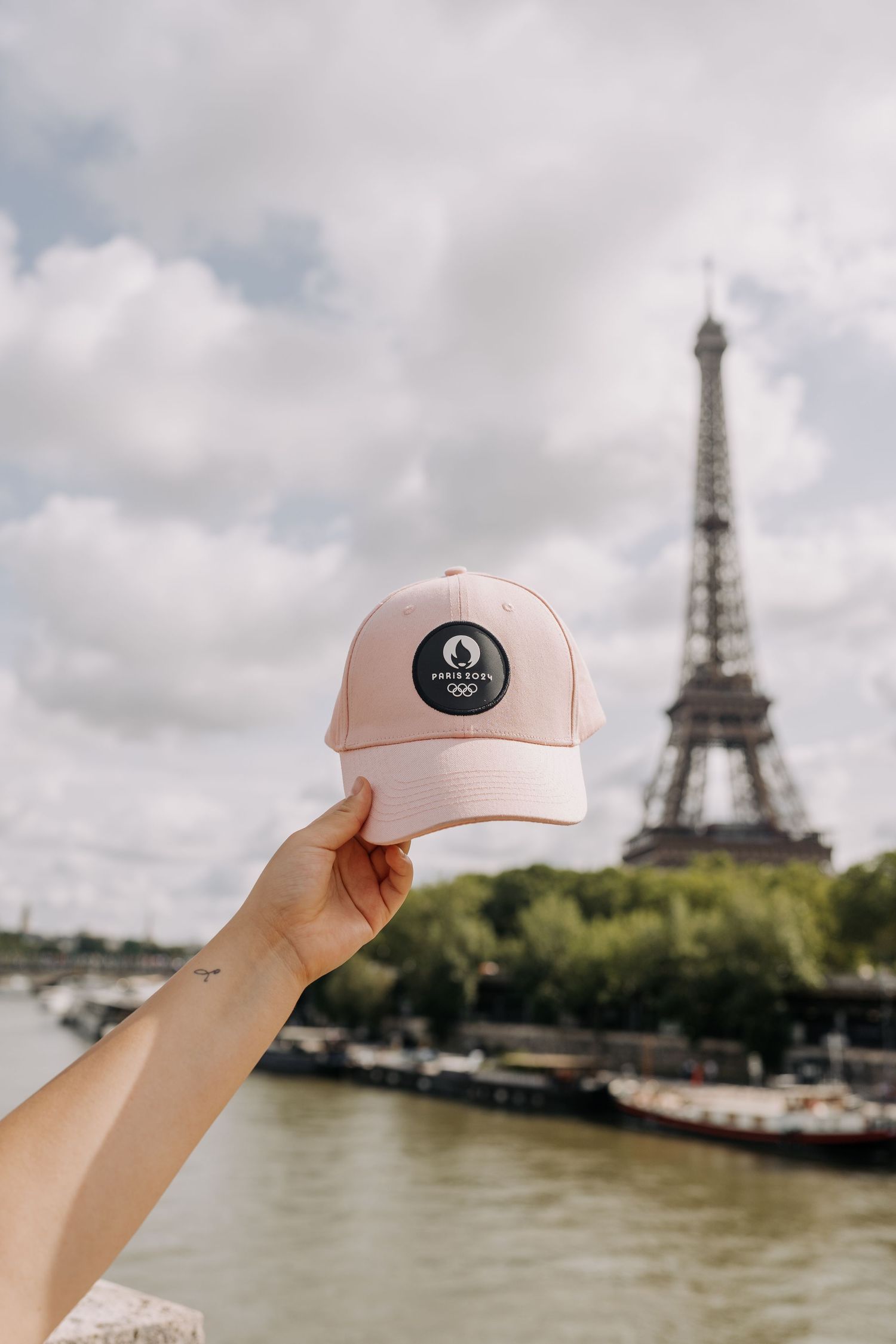 Hand holding pink Paris 2024 Olympics cap with Eiffel Tower in background on cloudy day.