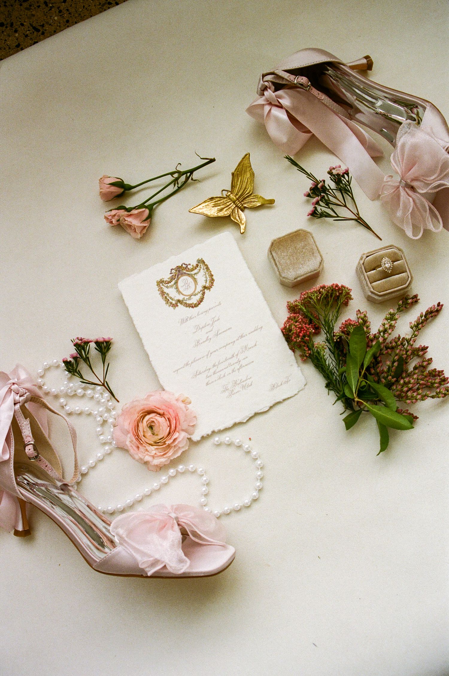 Romantic wedding flat lay with pink heels, pearl necklace, floral invitation, and gold butterfly.