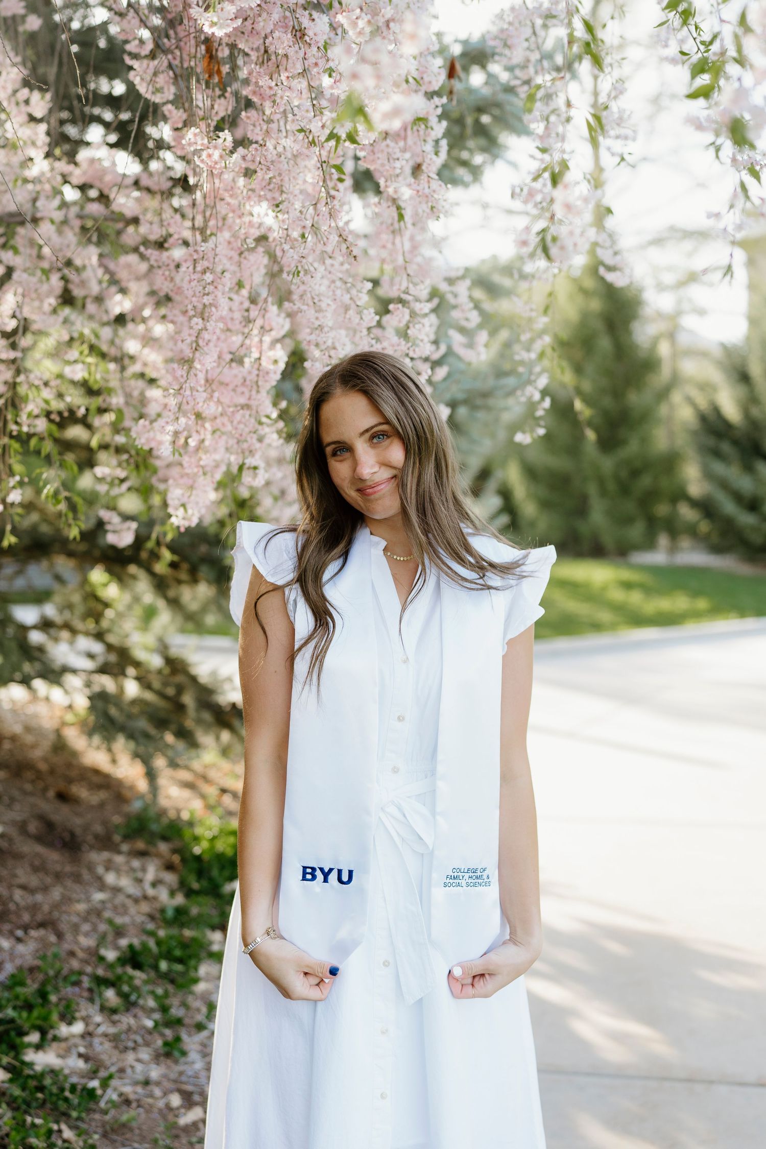 BYU graduate in white dress holding stole under pink cherry blossom tree.