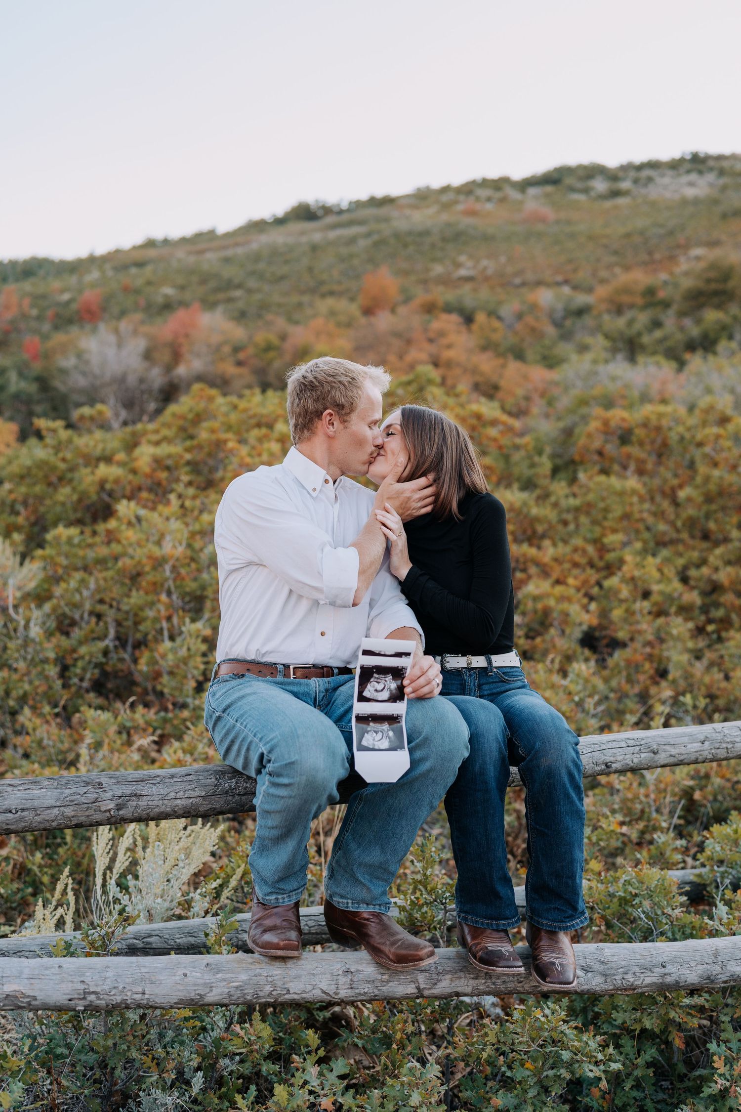 Couple kissing on wooden fence holding ultrasound photos during autumn pregnancy announcement.