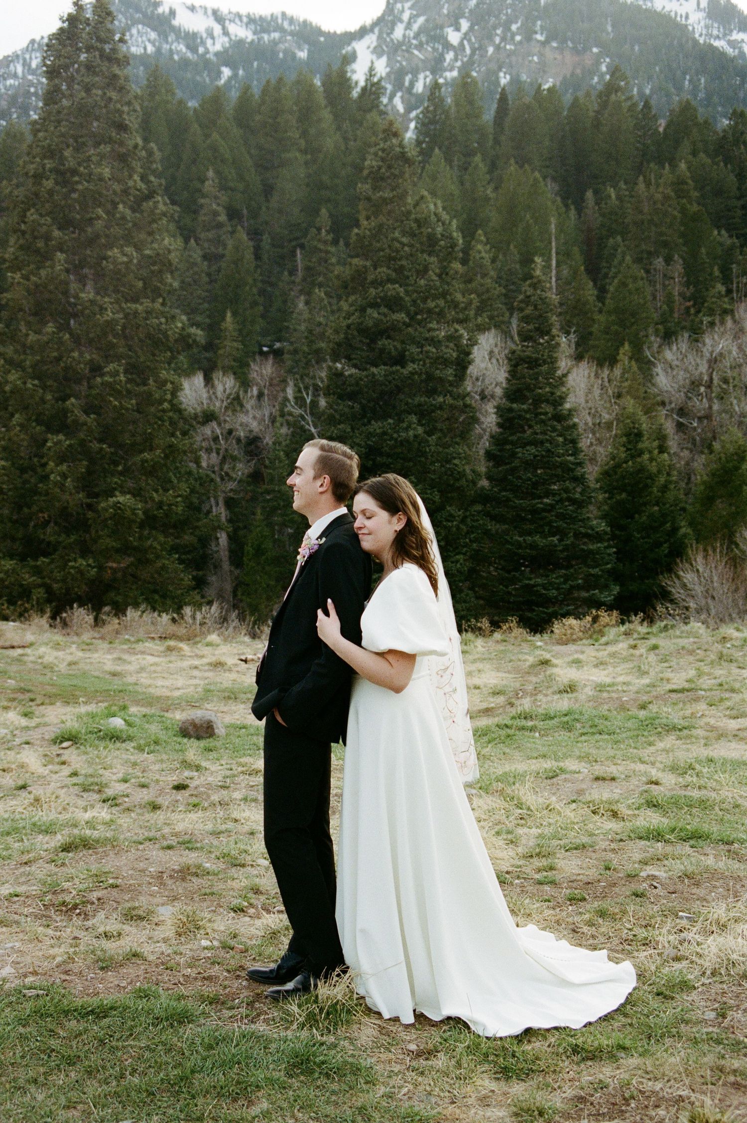 Bride and groom pose together in a mountain meadow, surrounded by tall evergreen trees and snow-capped peaks.