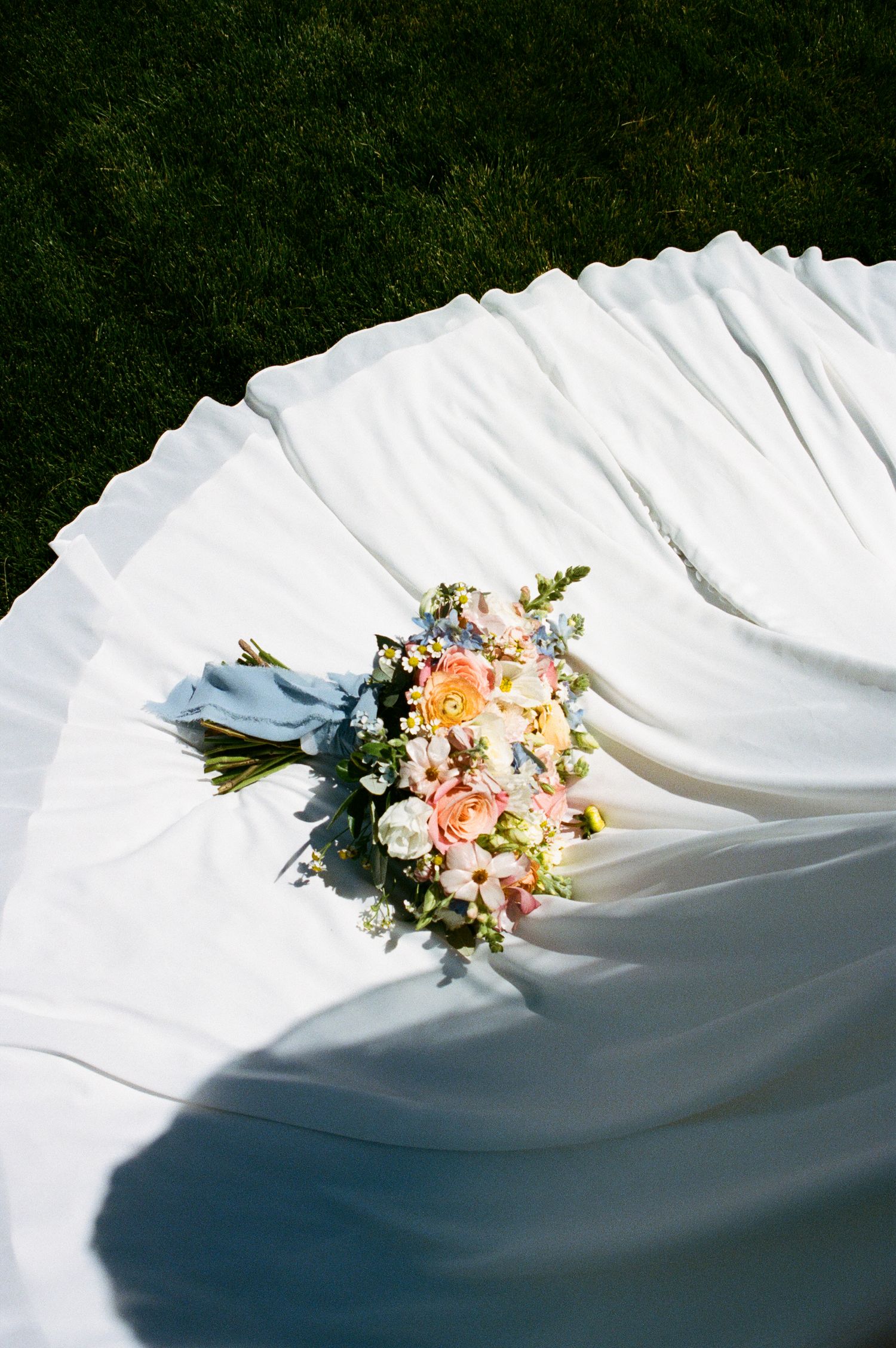 A colorful bridal bouquet of pink and white flowers rests on a flowing white wedding dress on grass.