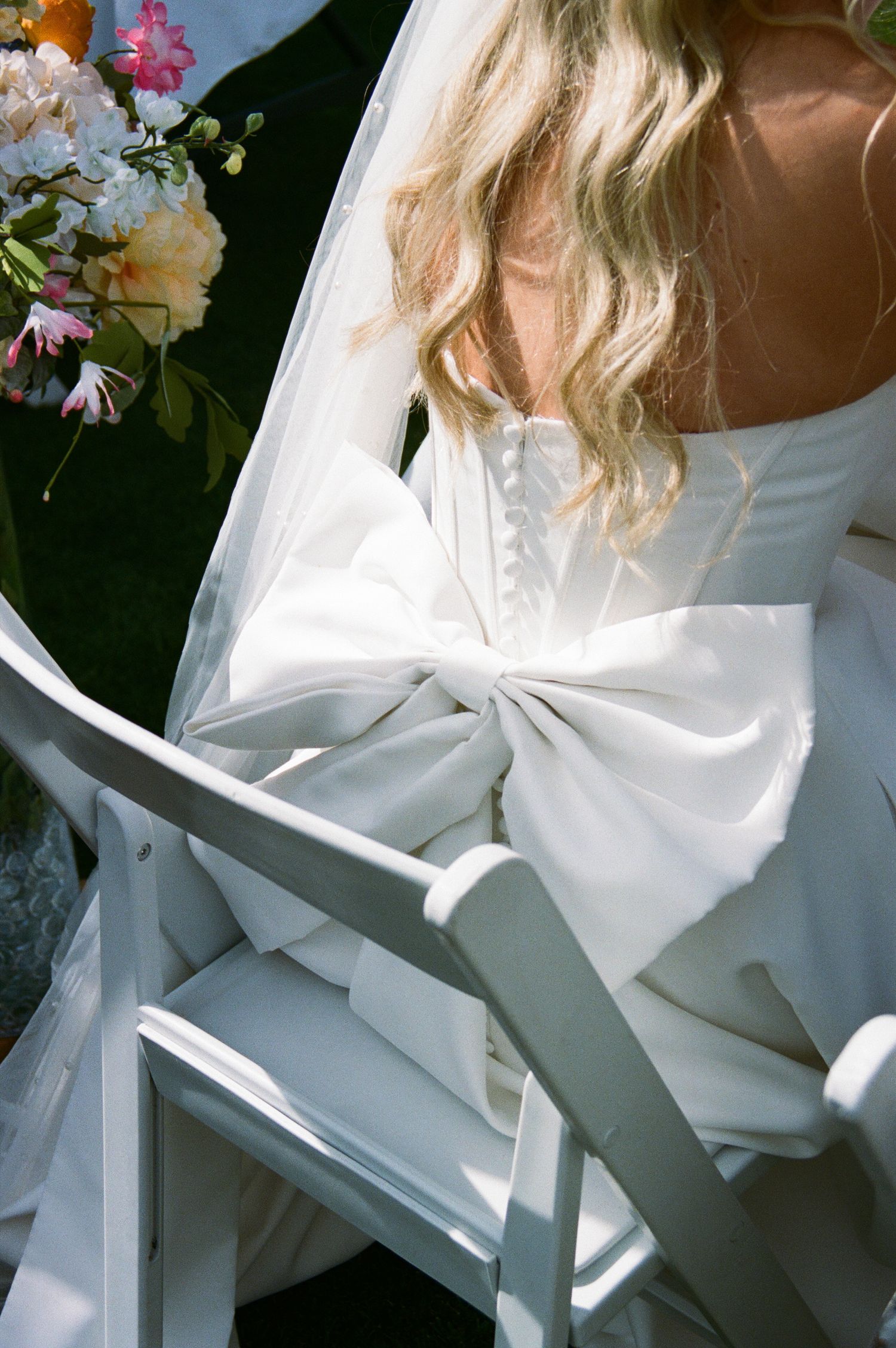 Close-up of bride in white wedding gown with large satin bow detail, seated outdoors in sunlight.