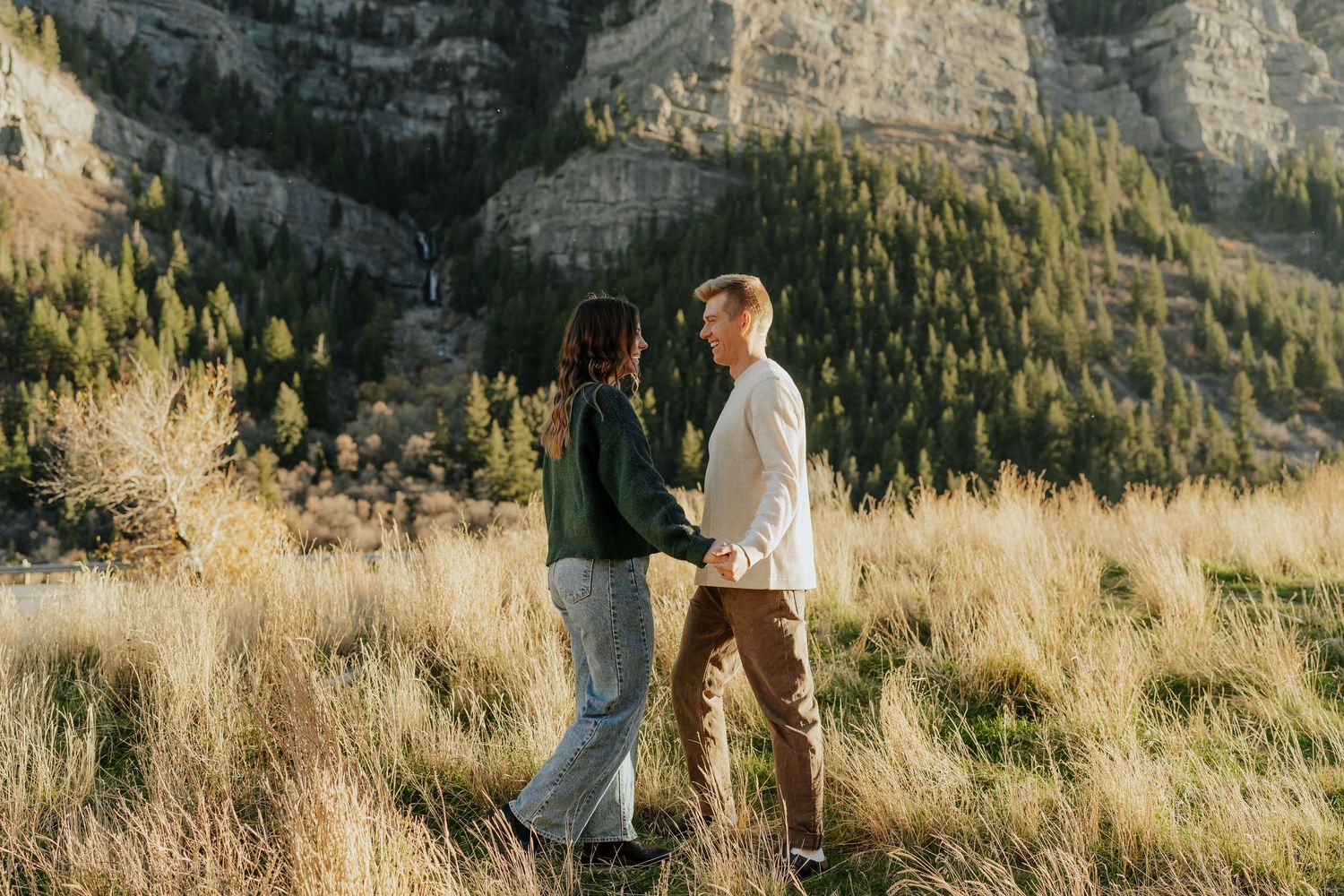 Couple holding hands and walking through tall golden grass with mountain scenery in background in Provo, Utah.
