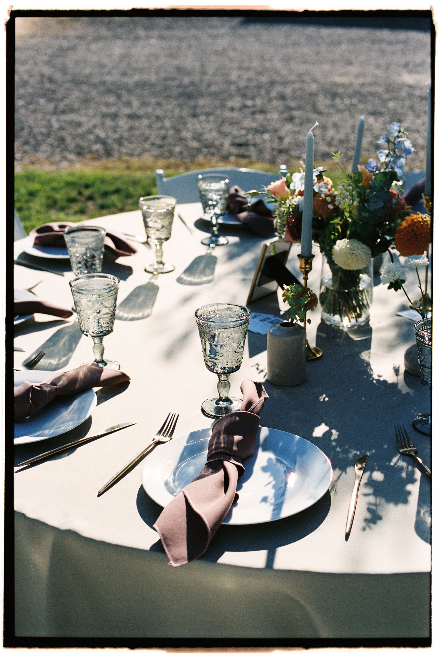 Elegant outdoor wedding table setting with crystal goblets, mauve napkins, gold cutlery, and a floral centerpiece on a white linen.