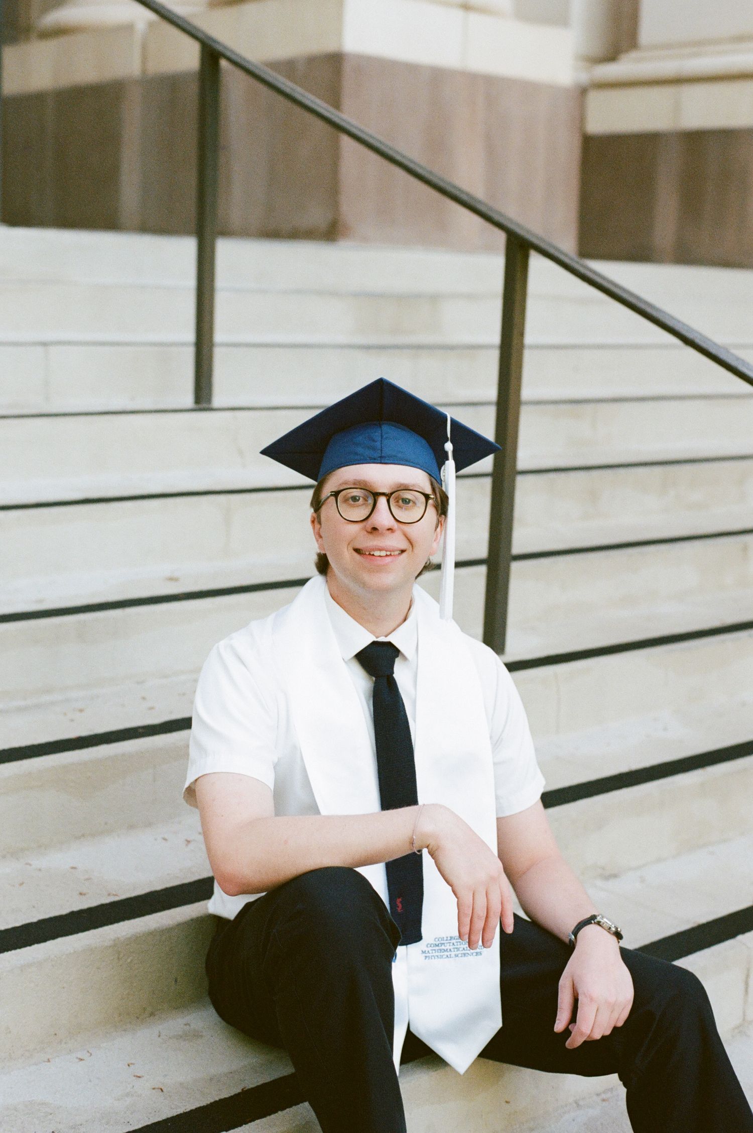 Smiling graduate in cap and glasses sits on stone steps holding diploma, wearing white shirt and black tie.