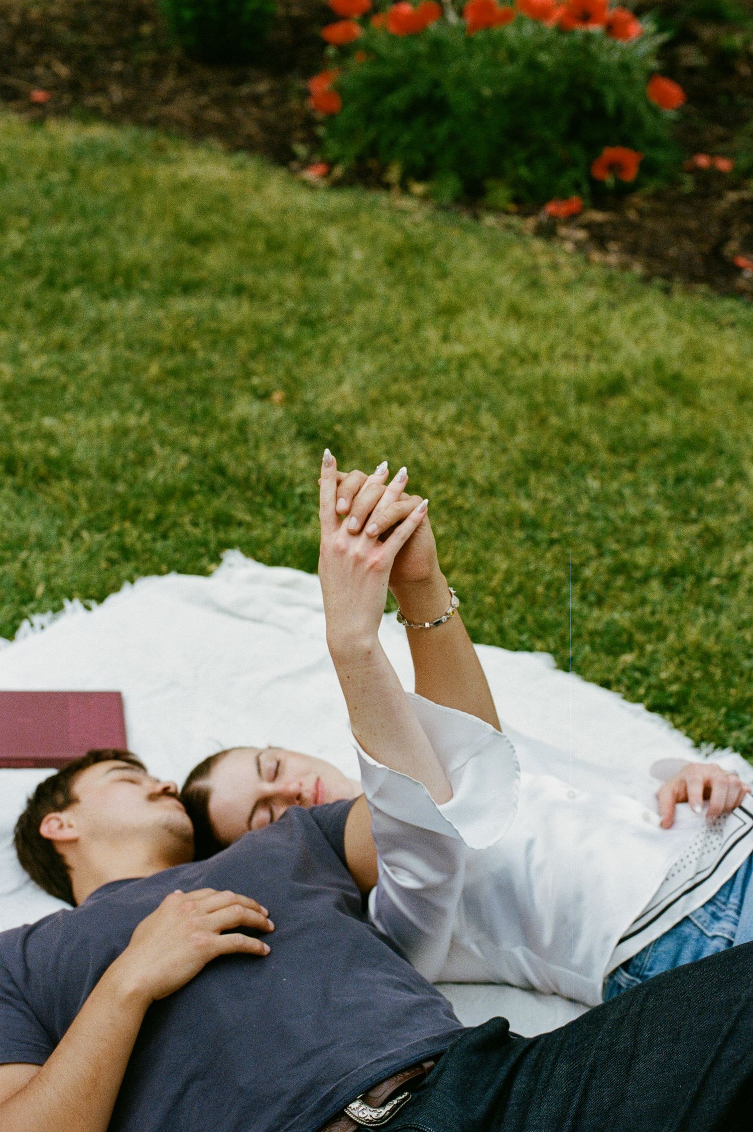 Couple lying on white blanket in grass, holding hands up, with red flowers in background.