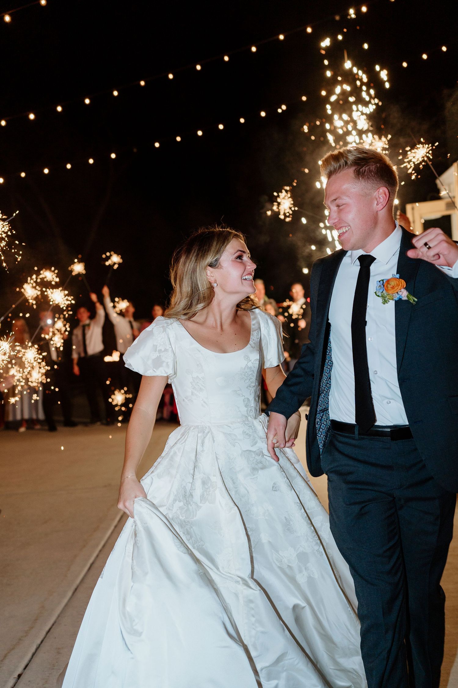 Bride and groom walk through sparkler exit at night with string lights glowing in the background.