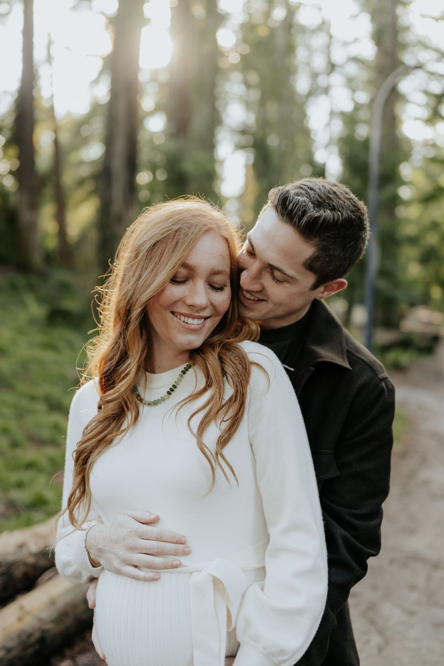 Man embracing pregnant woman from behind in forest, both smiling warmly in golden sunlight in Davis, California.