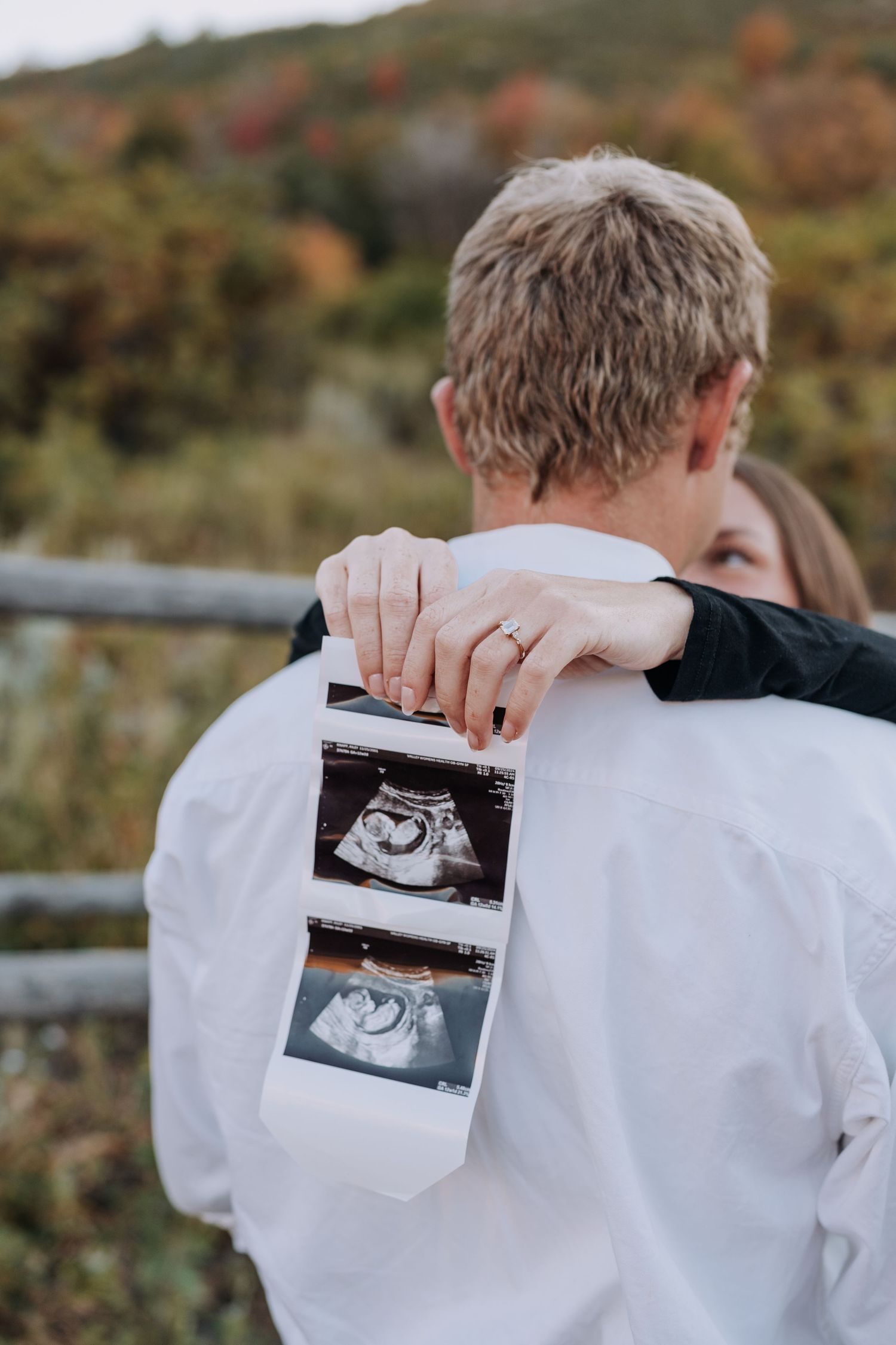 Woman hugging man from behind holding ultrasound photos during outdoor fall pregnancy reveal.