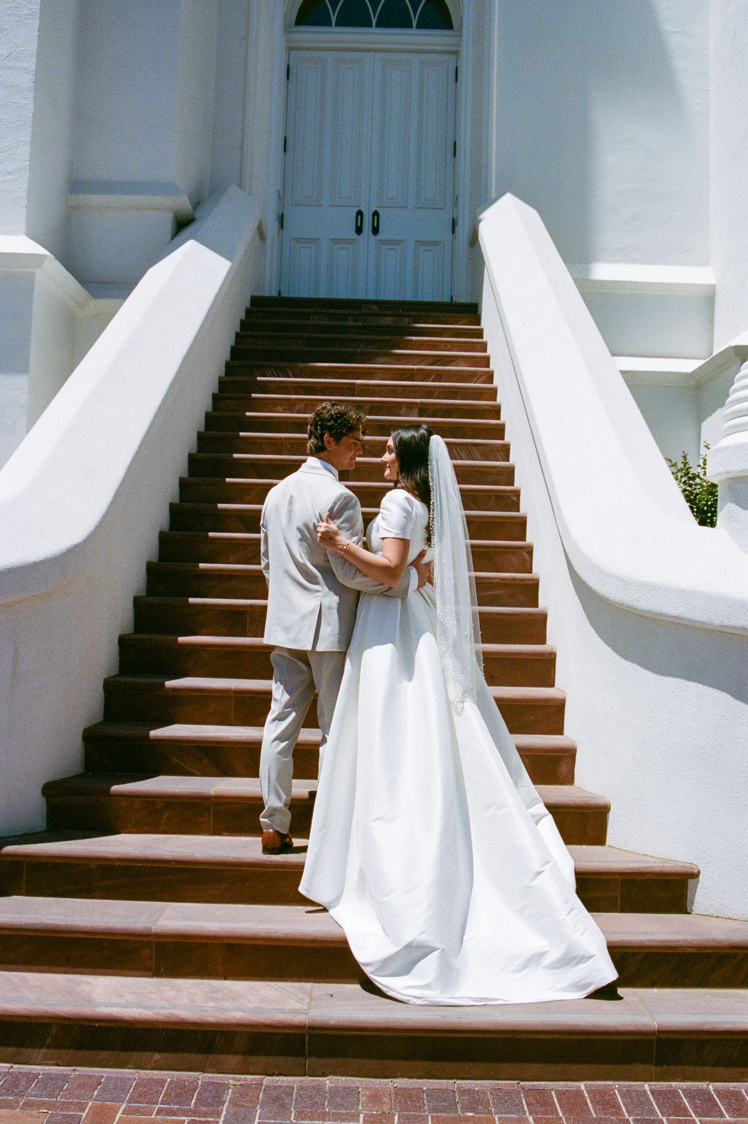 Bride and groom embrace on brick stairs outside a white church on their wedding day.