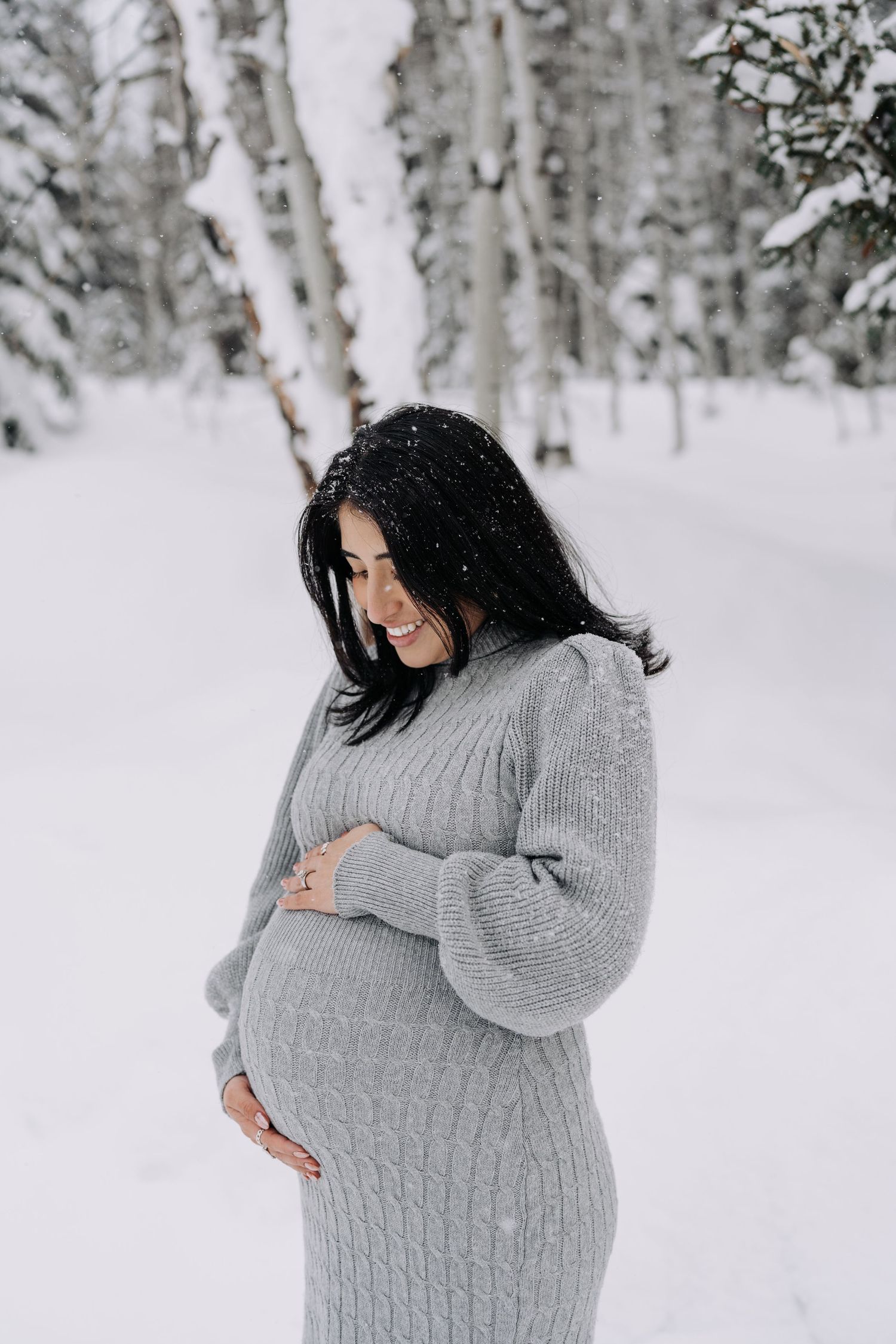 Smiling pregnant woman in gray knit dress cradling baby bump in snowy winter forest.