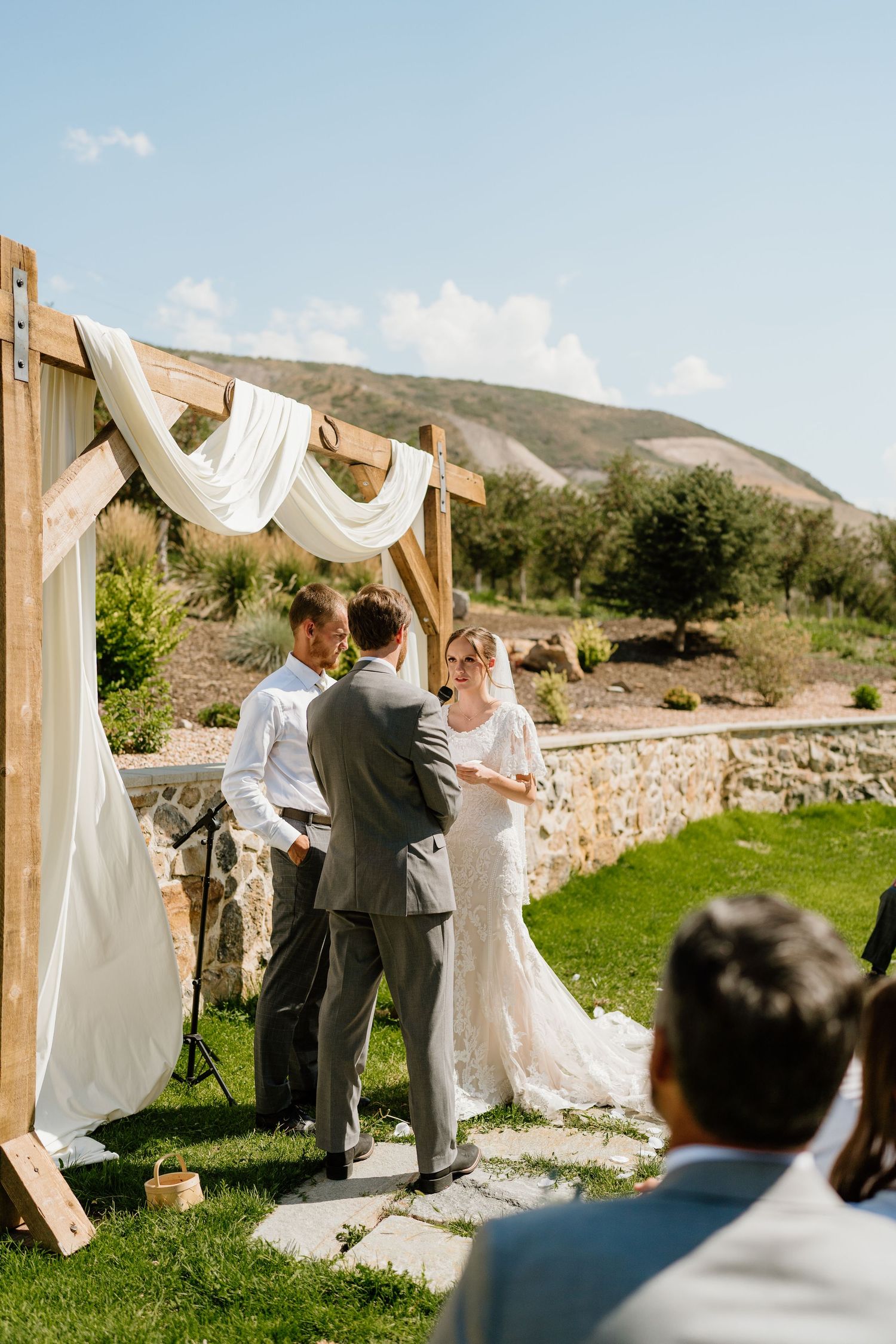 Bride and groom exchange vows outdoors under wooden arch draped with white fabric near stone wall.