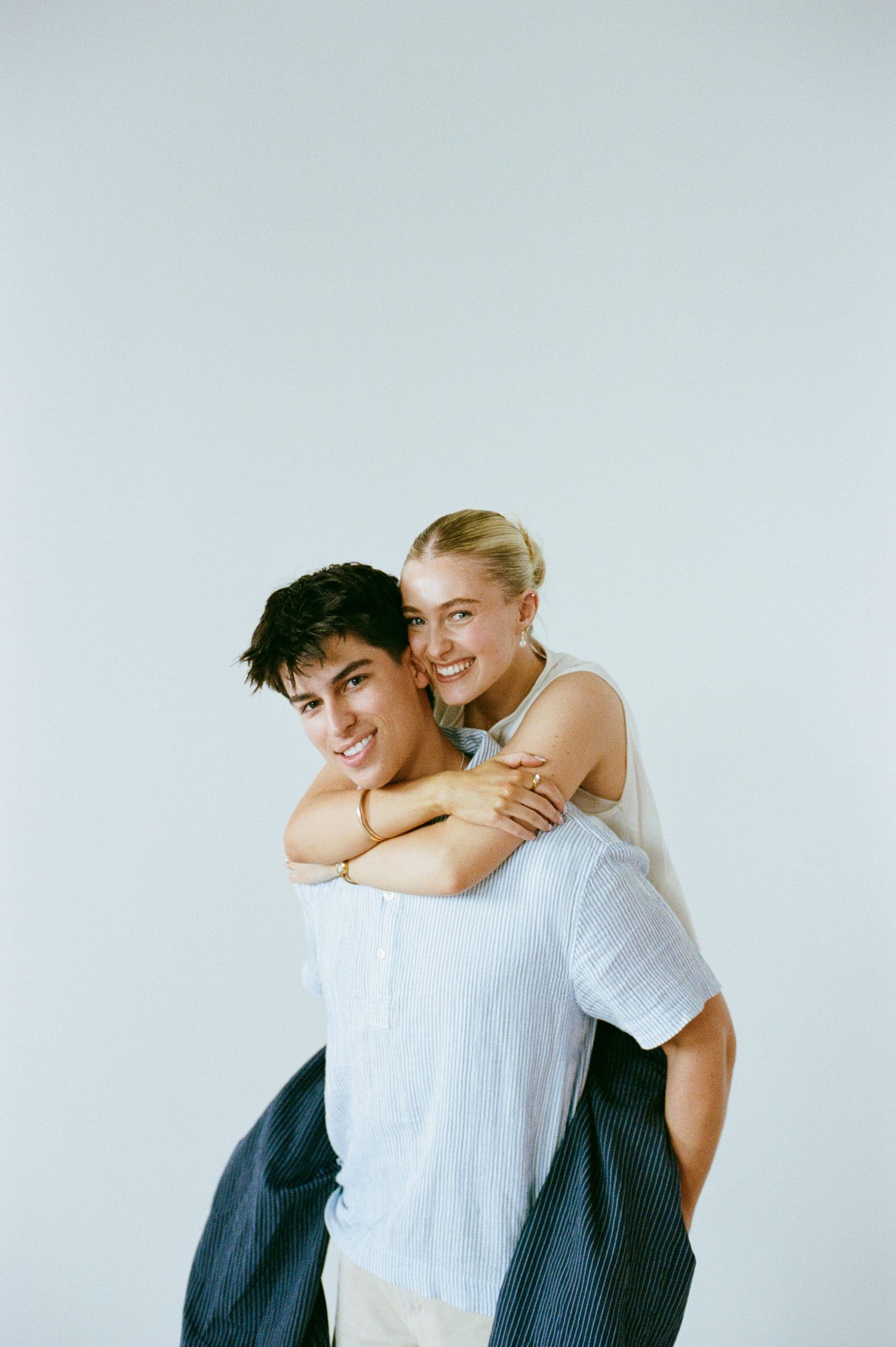 Smiling young man giving piggyback ride to happy blonde woman against light gray background.