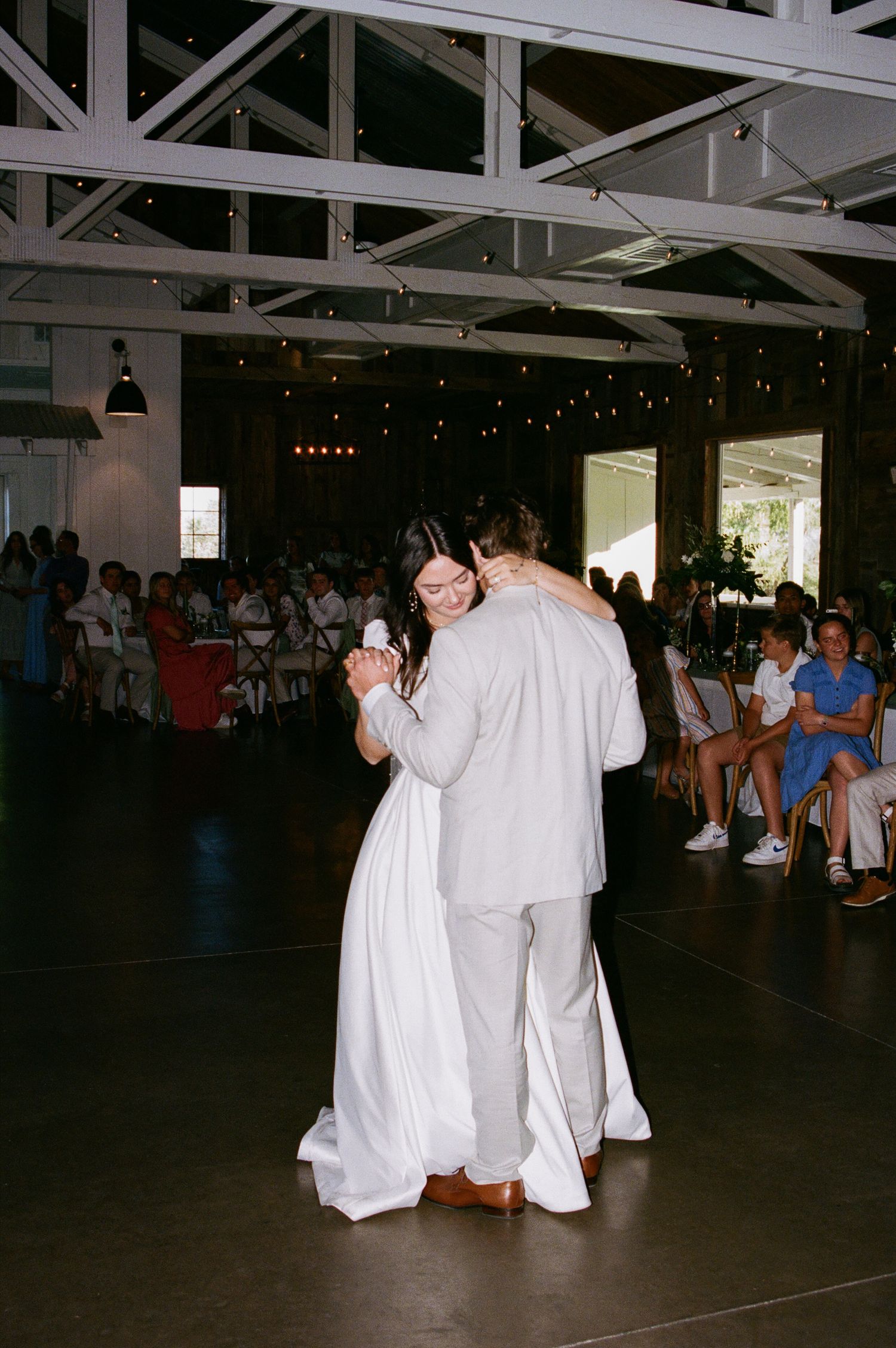Bride and groom sharing first dance in rustic barn venue with string lights and seated guests.