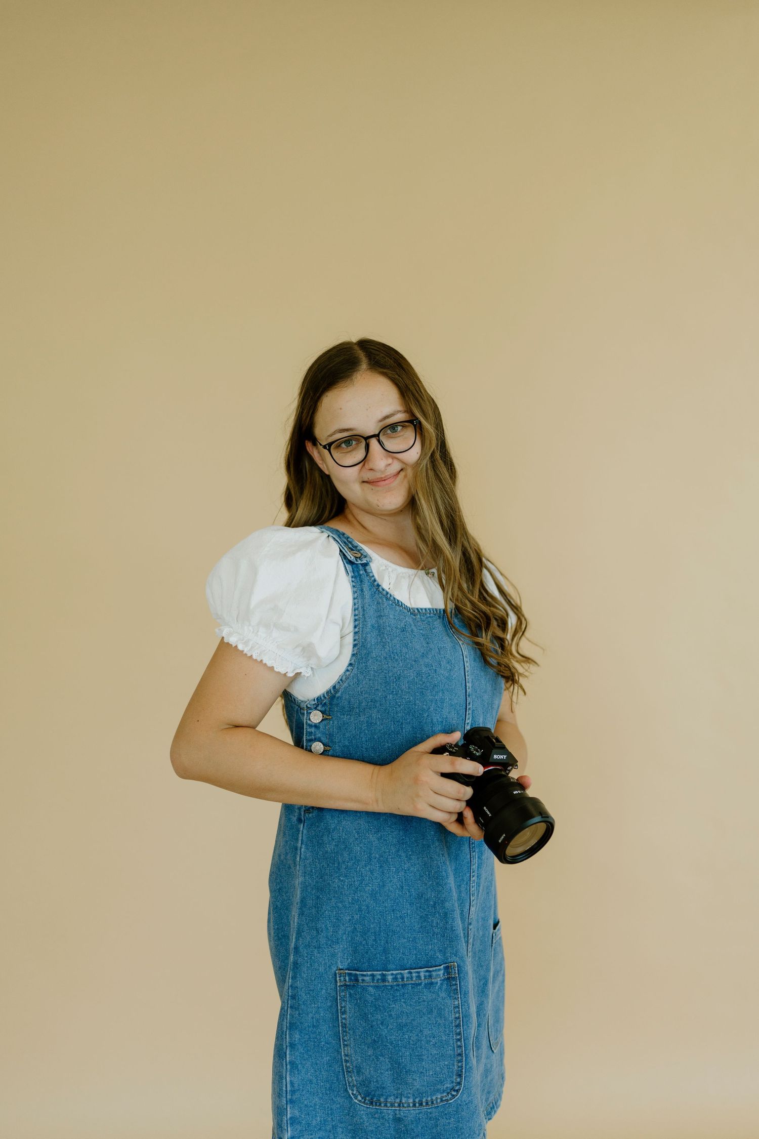 Young female photographer smiling in denim overalls holding a DSLR camera against a beige background.