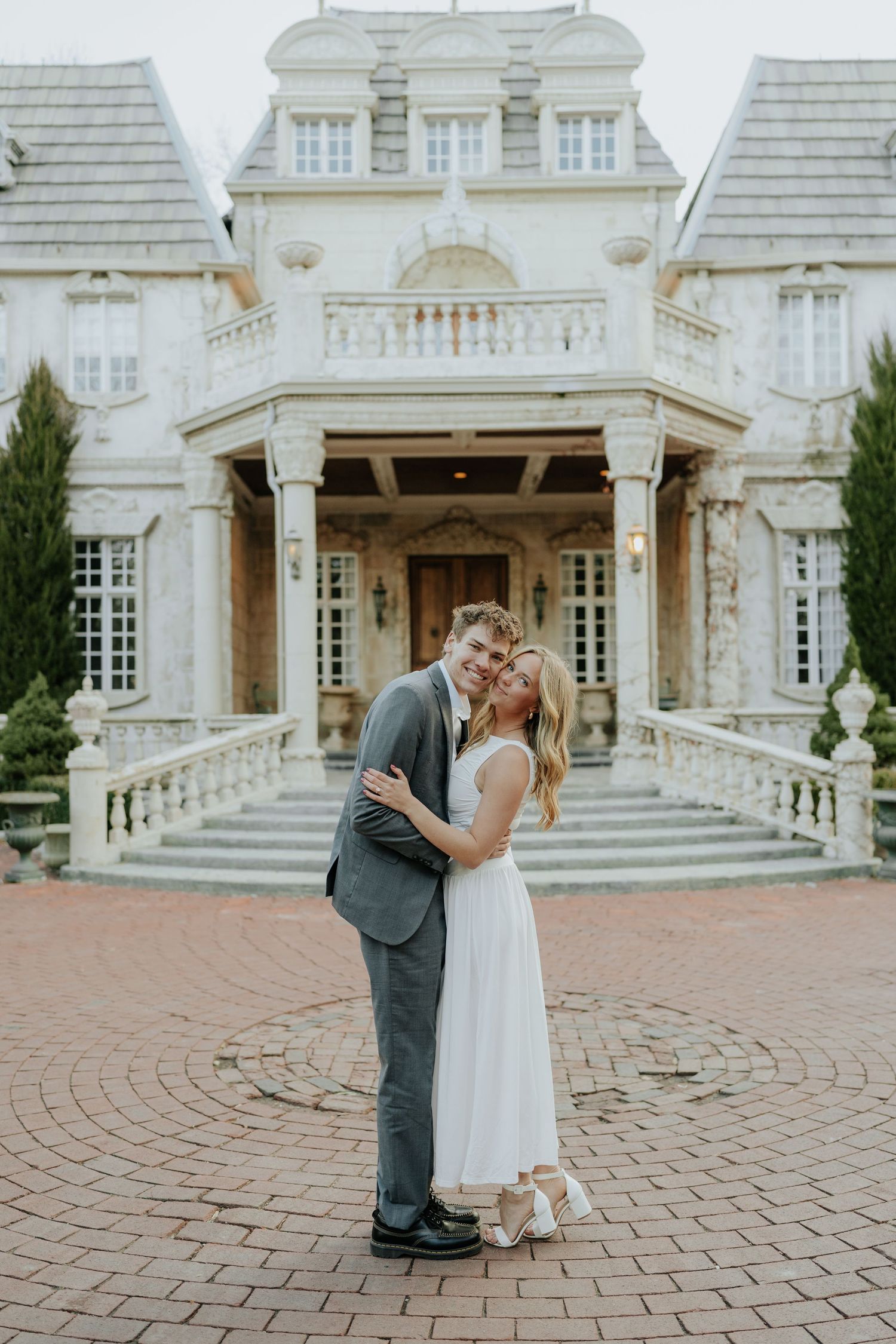 Couple in wedding attire embracing in front of an elegant stone mansion with grand staircase at La Caille Restaurant in Utah.