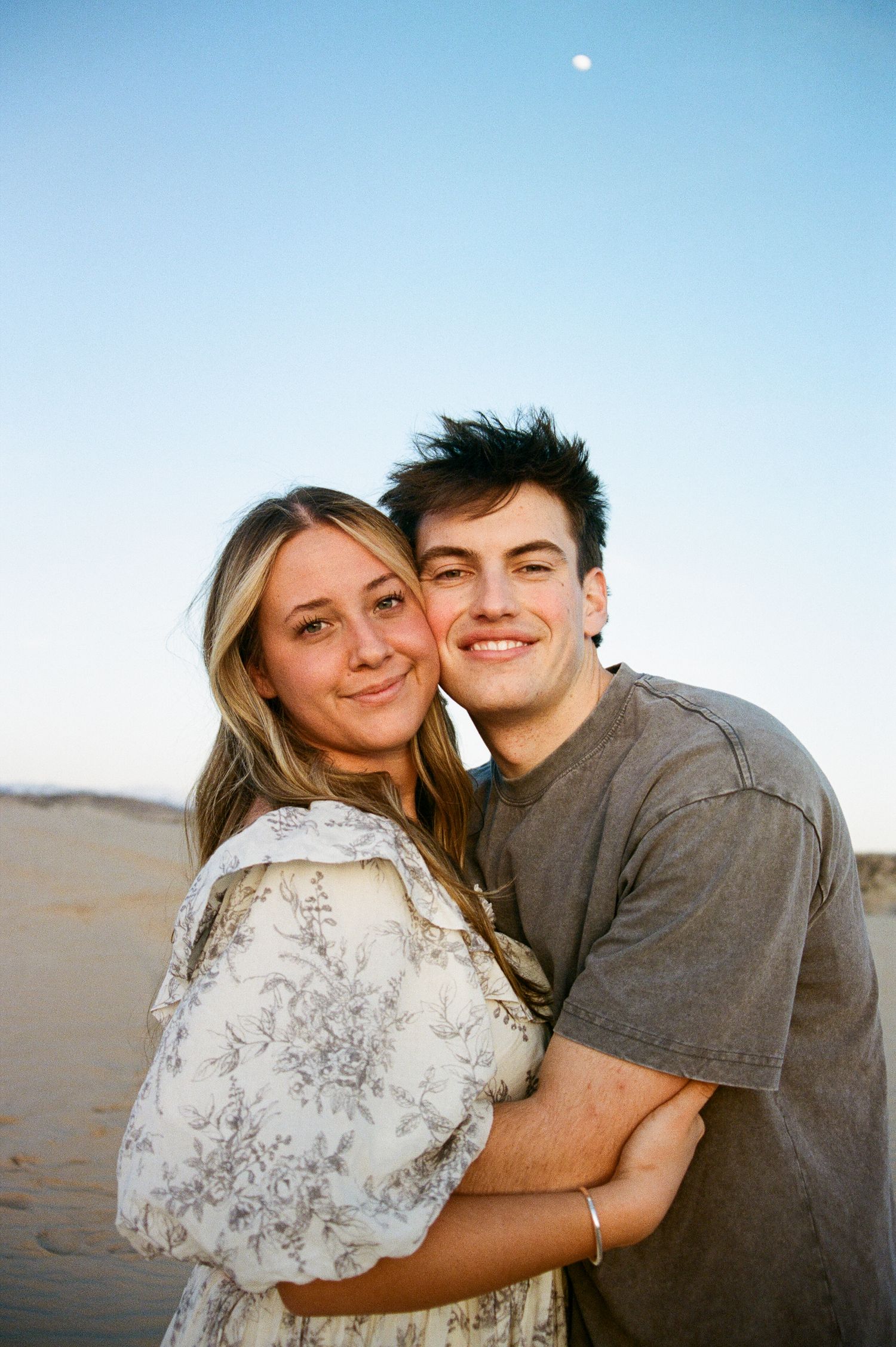 Smiling young couple embraces on sandy dunes at dusk with a clear blue sky and moon above.