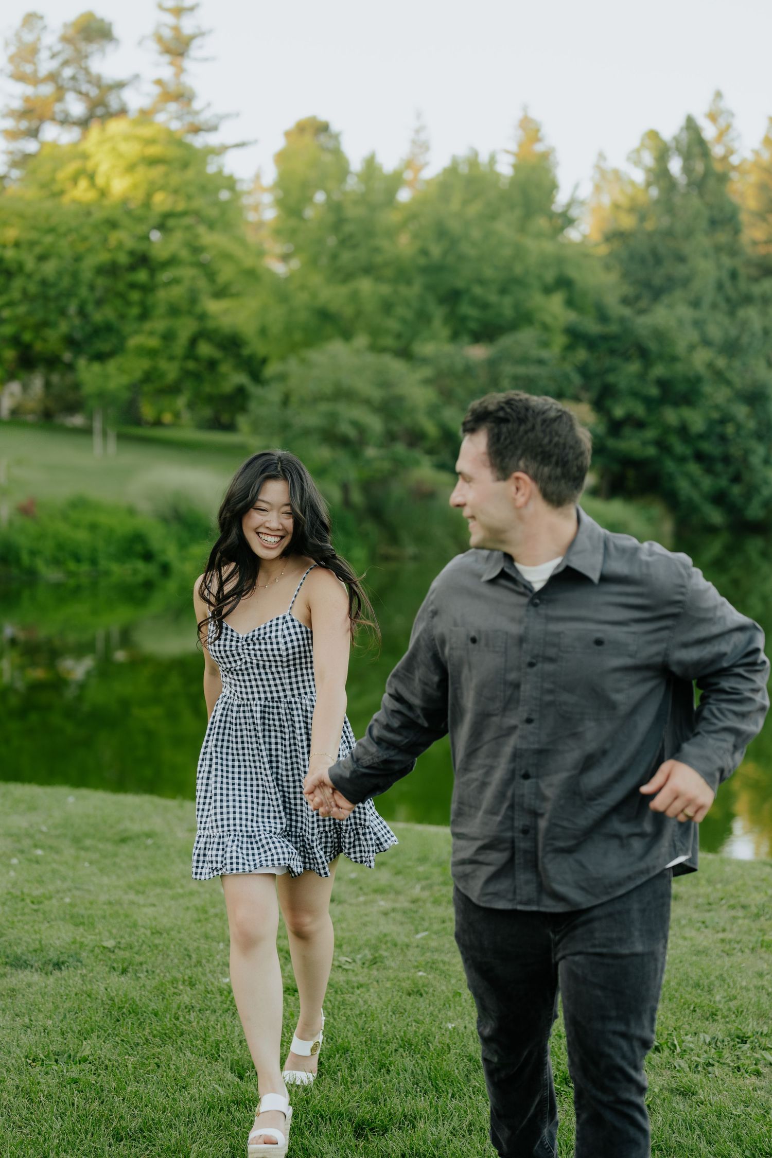 A smiling couple holds hands while walking on green grass near a pond surrounded by lush trees at dusk.