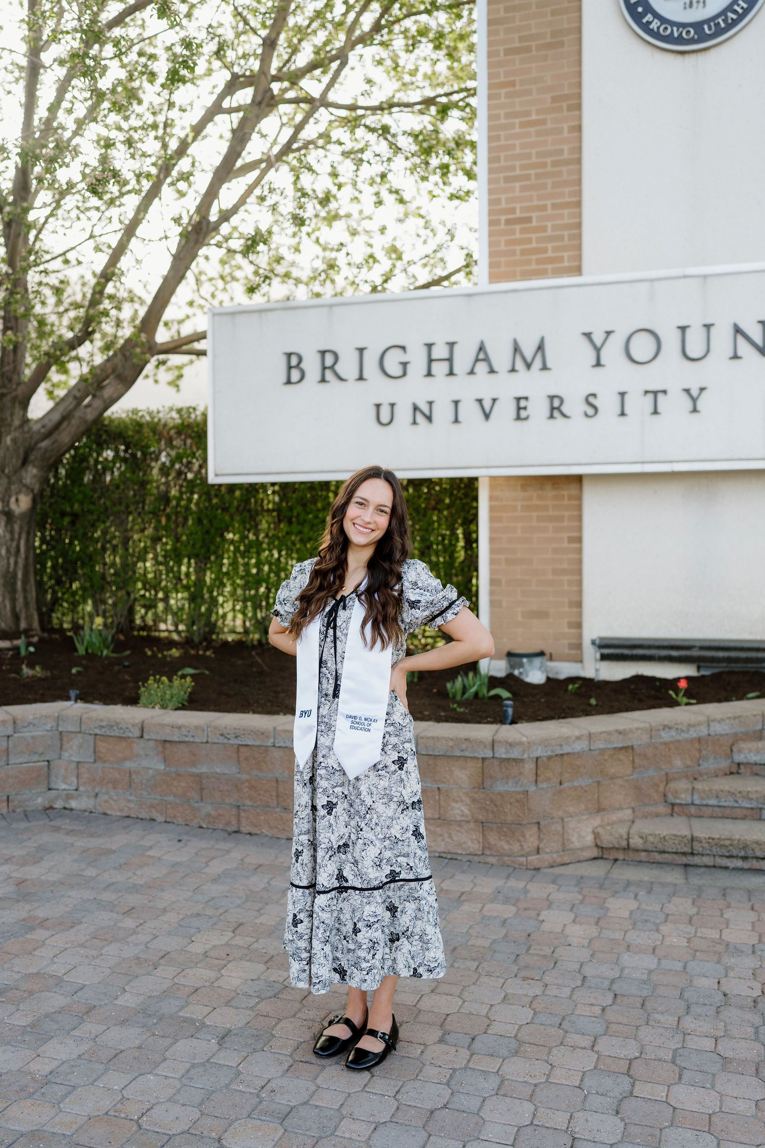 Smiling BYU graduate in floral dress standing in front of Brigham Young University sign.