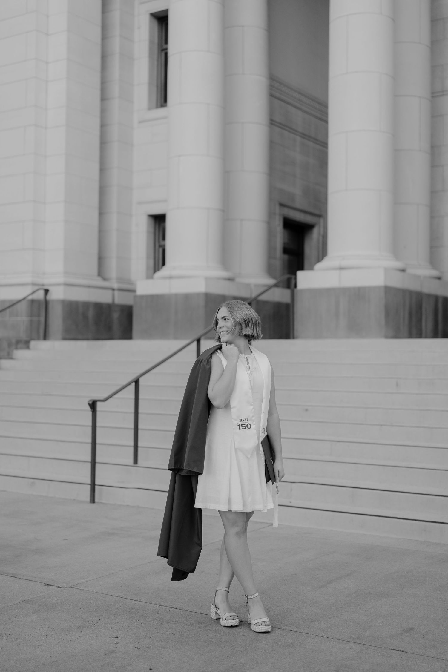 Black and white photo of a woman in a white dress holding a graduation gown on building steps.
