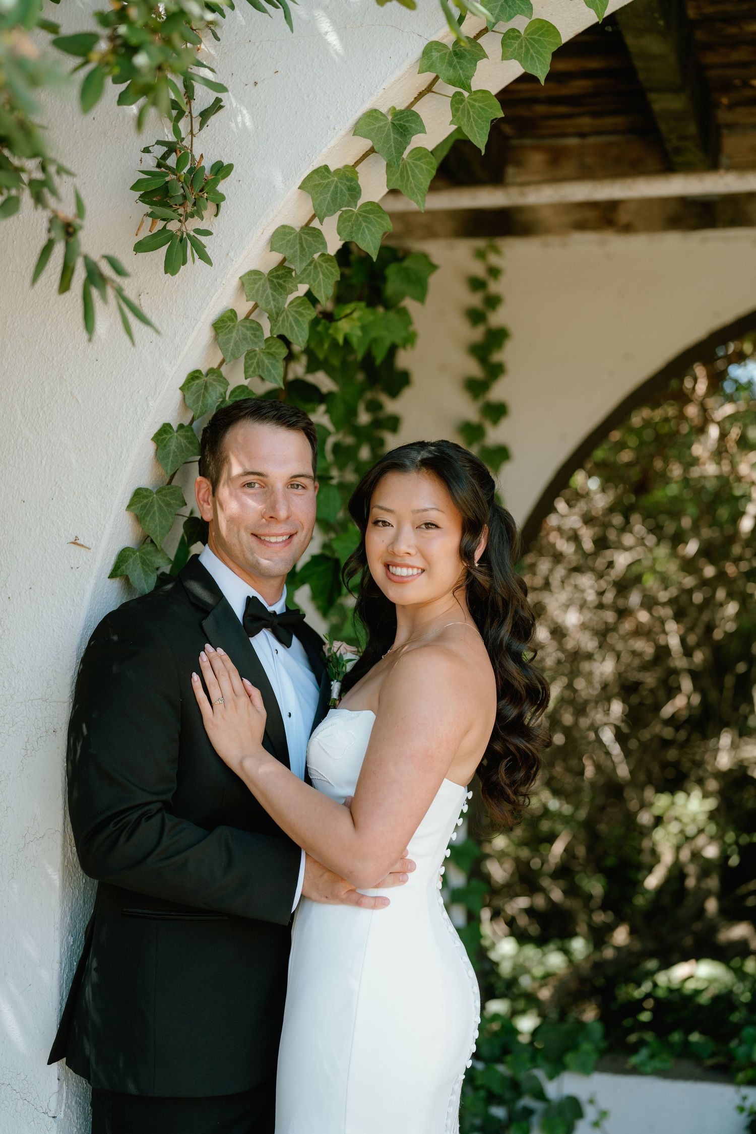 Bride and groom pose together in front of ivy-covered white arch, she in strapless gown, he in black tuxedo.