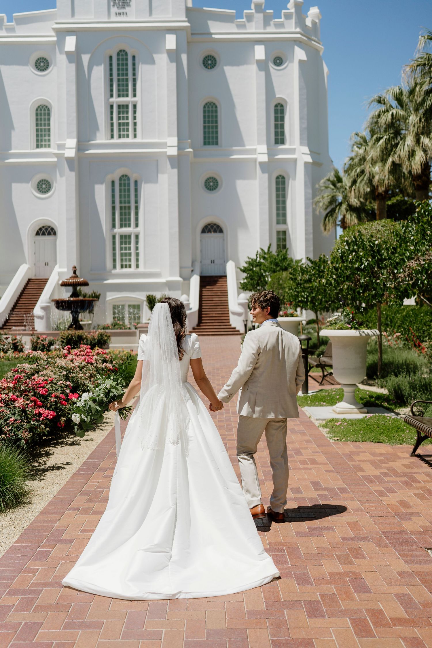 Bride and groom walk hand in hand toward a grand white temple on their sunny wedding day in Saint George, Utah