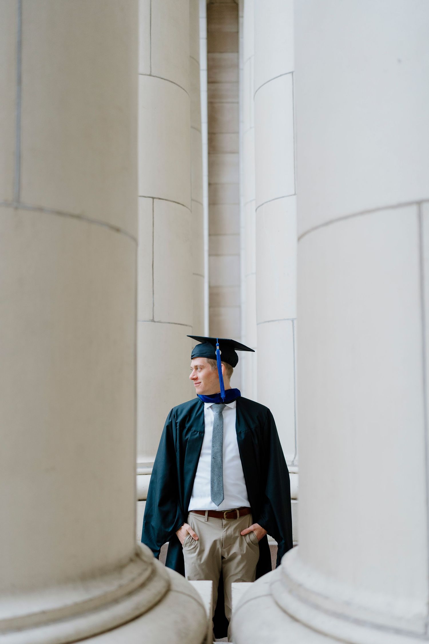 Male graduate in dark green gown standing between tall stone columns looking away.