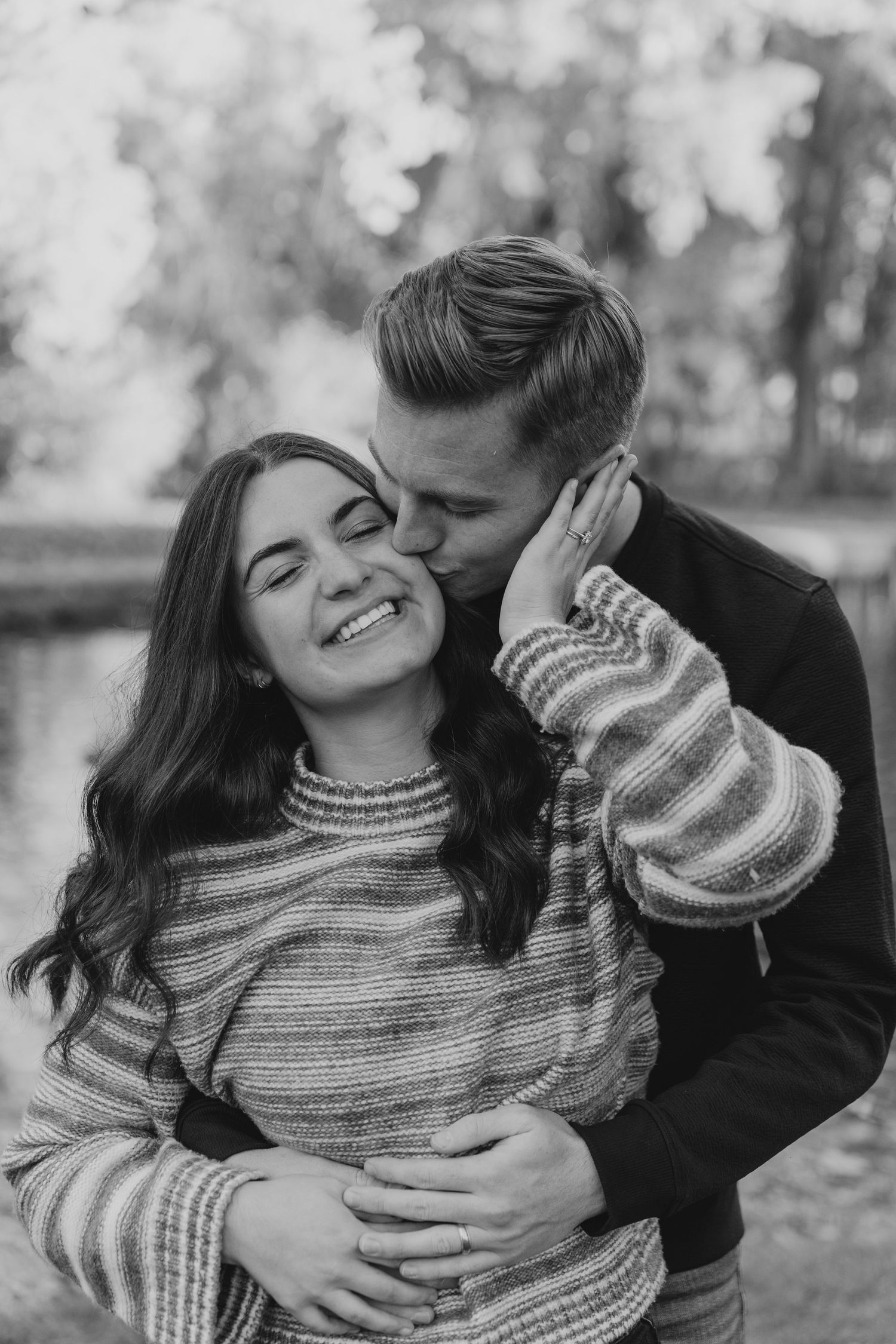 Black and white photo of man kissing smiling woman on cheek during fall engagement session.