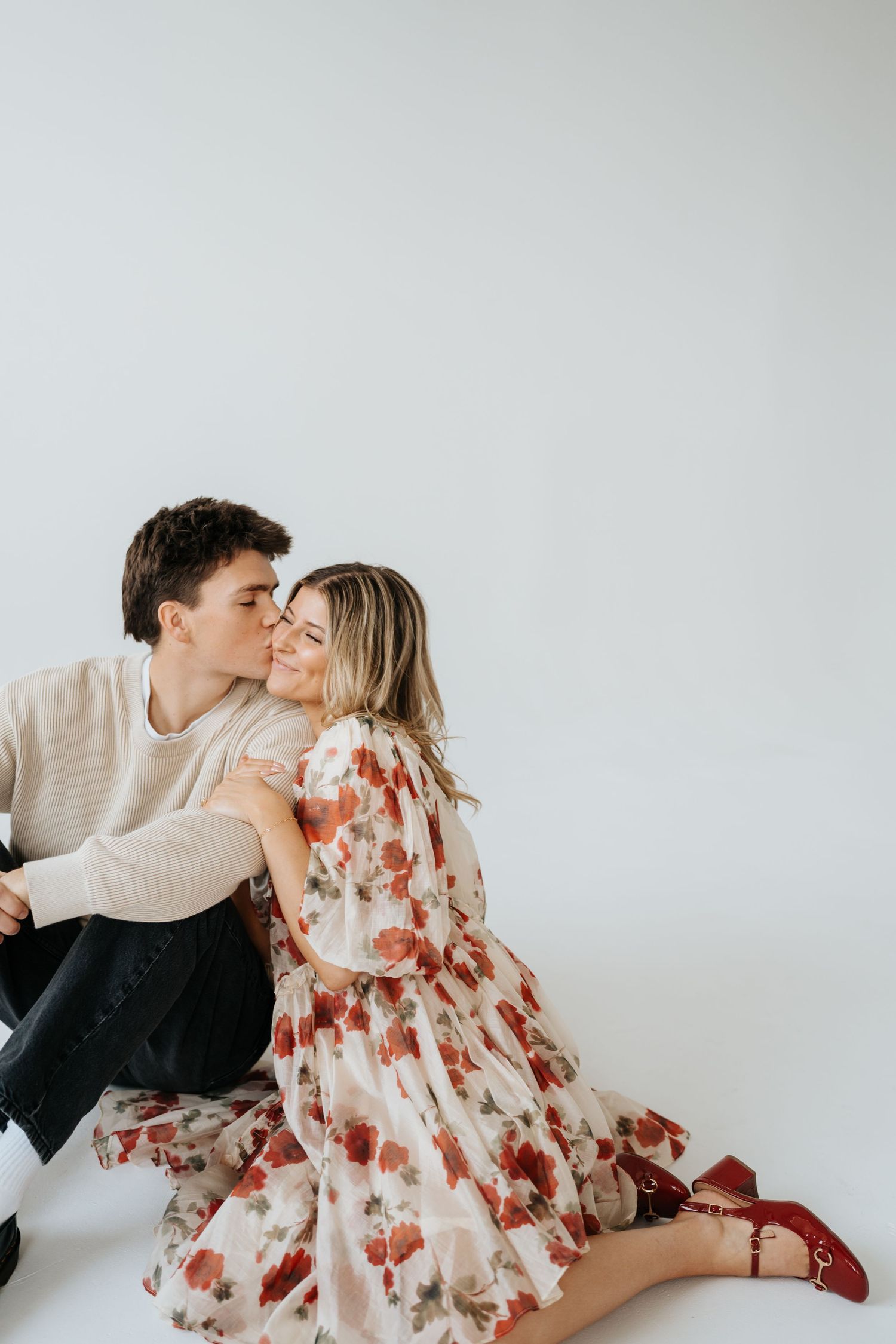 Couple sitting on white studio floor, man kissing woman's cheek as she smiles in floral dress at Studio Elevn in Salt Lake City, Utah.