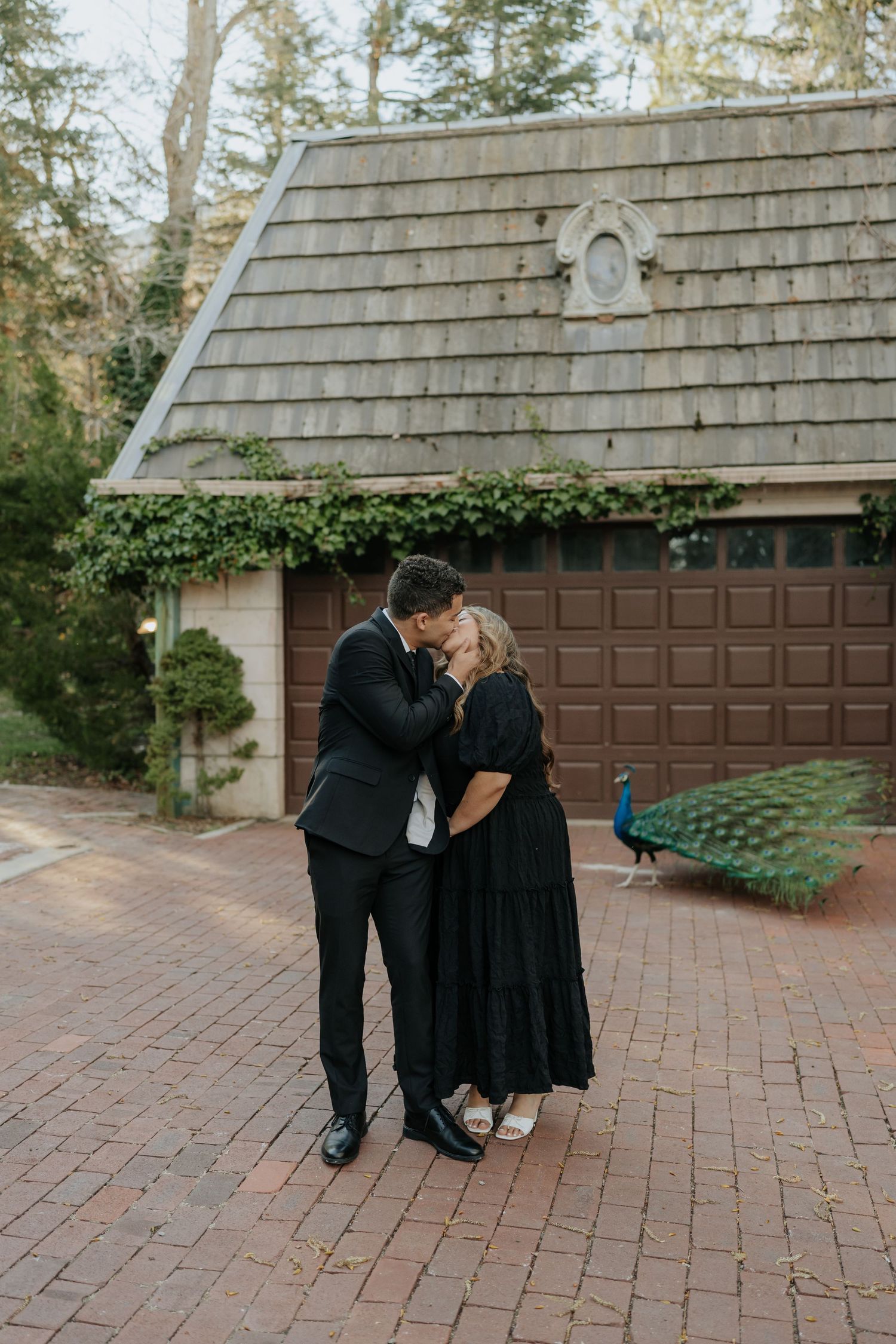 Couple in matching black outfits sharing a kiss on a brick driveway with a peacock nearby at La Caille Restaurant in Utah.