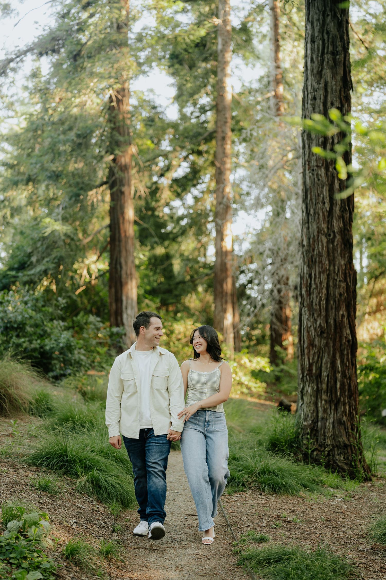Couple holding hands and laughing while walking on a forest path surrounded by tall redwood trees.
