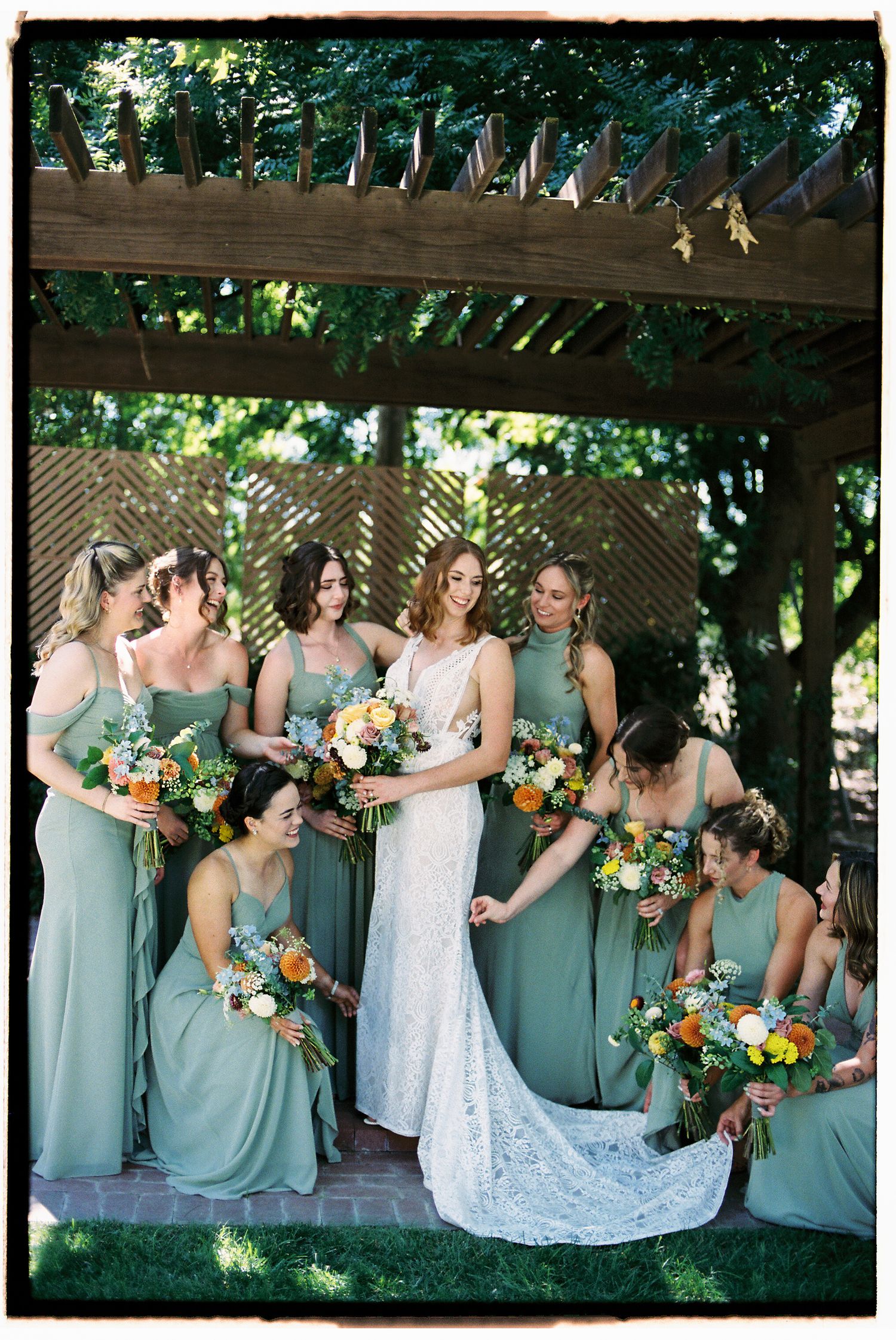Bride in white lace gown surrounded by bridesmaids in sage green dresses holding colorful bouquets under a garden pergola.