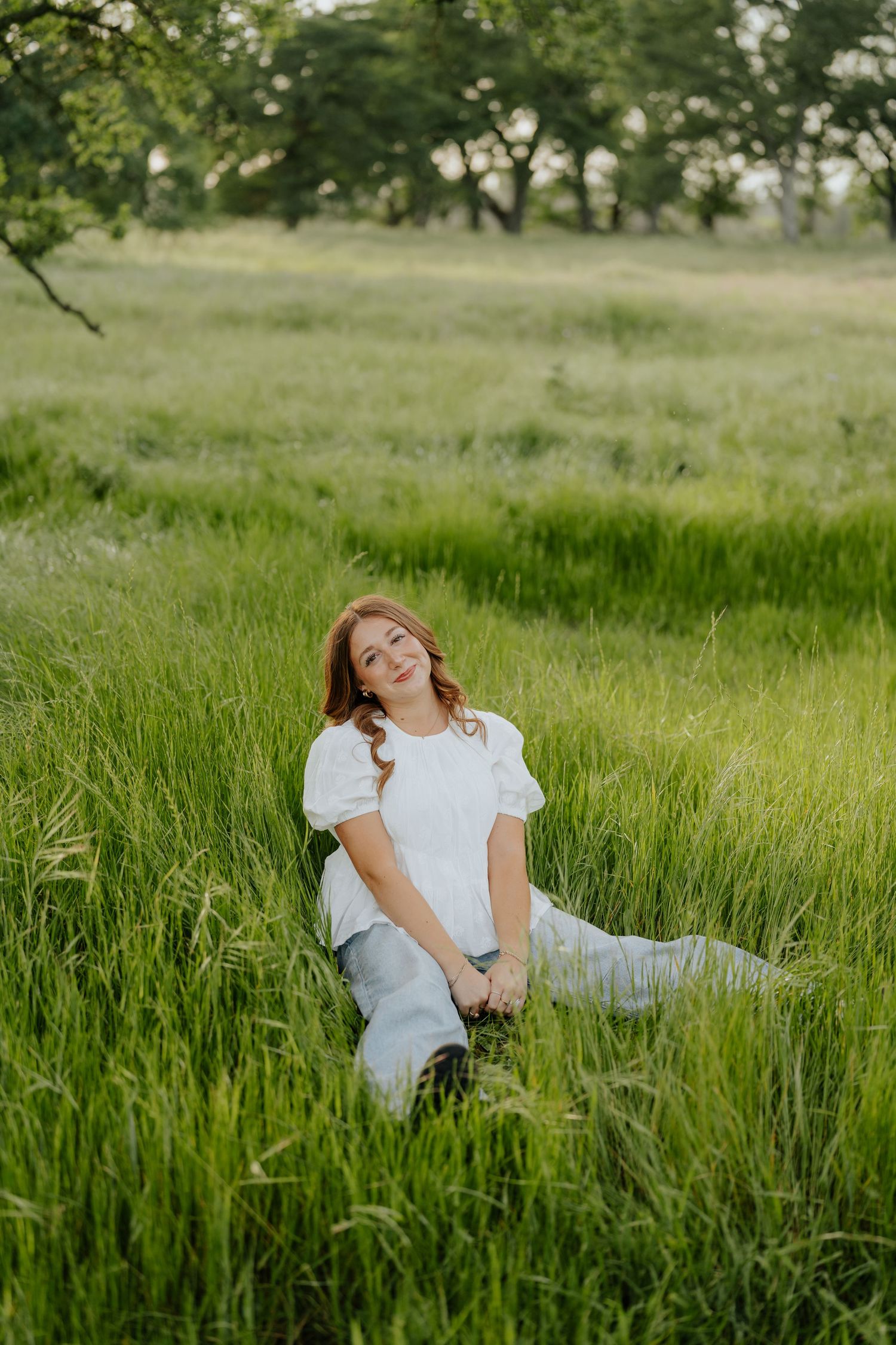 Young woman in white top and jeans sitting relaxed in lush green meadow grass with trees in background.