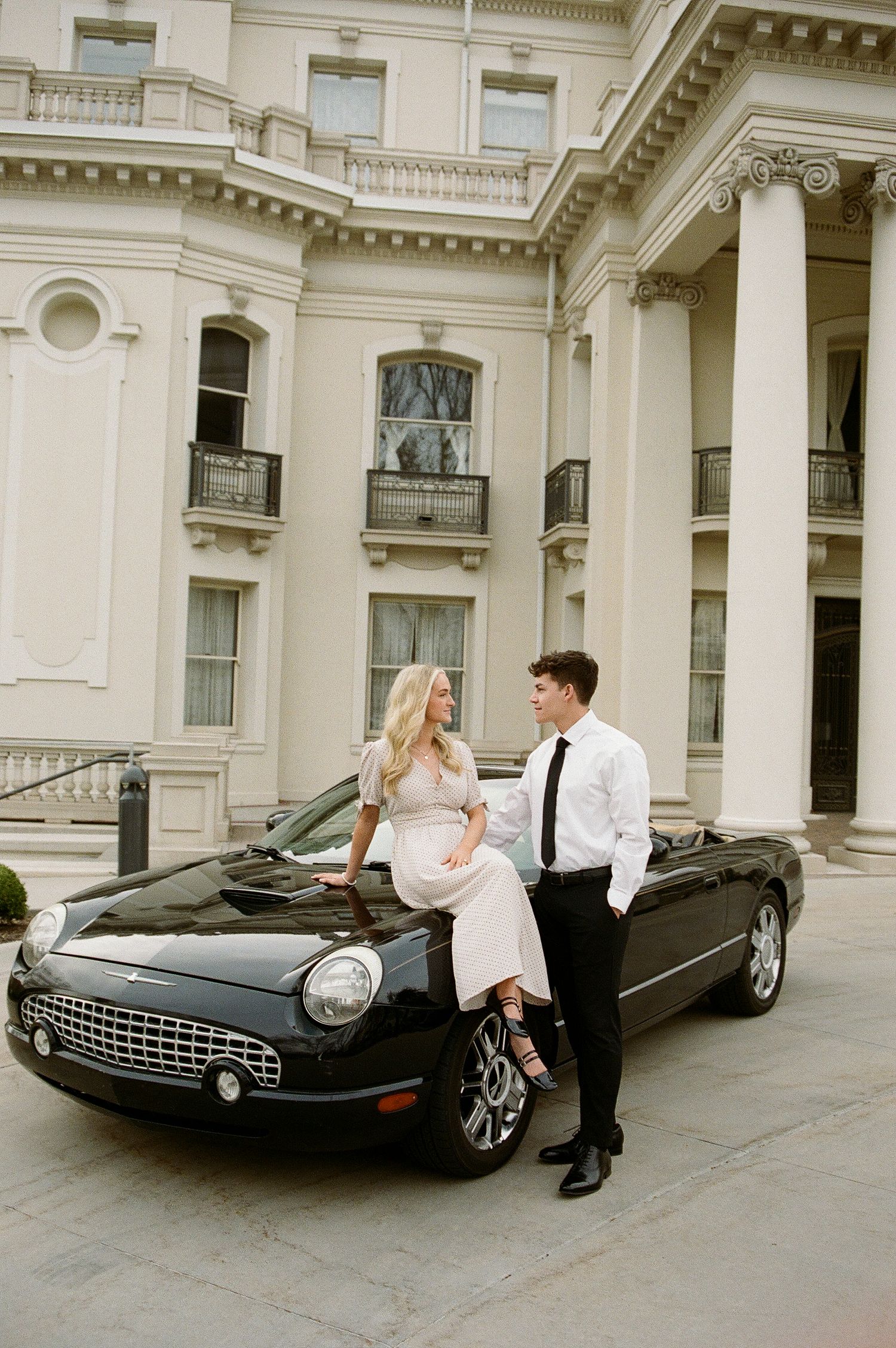 Elegant couple poses beside a black Ford Thunderbird convertible in front of a grand white neoclassical mansion.