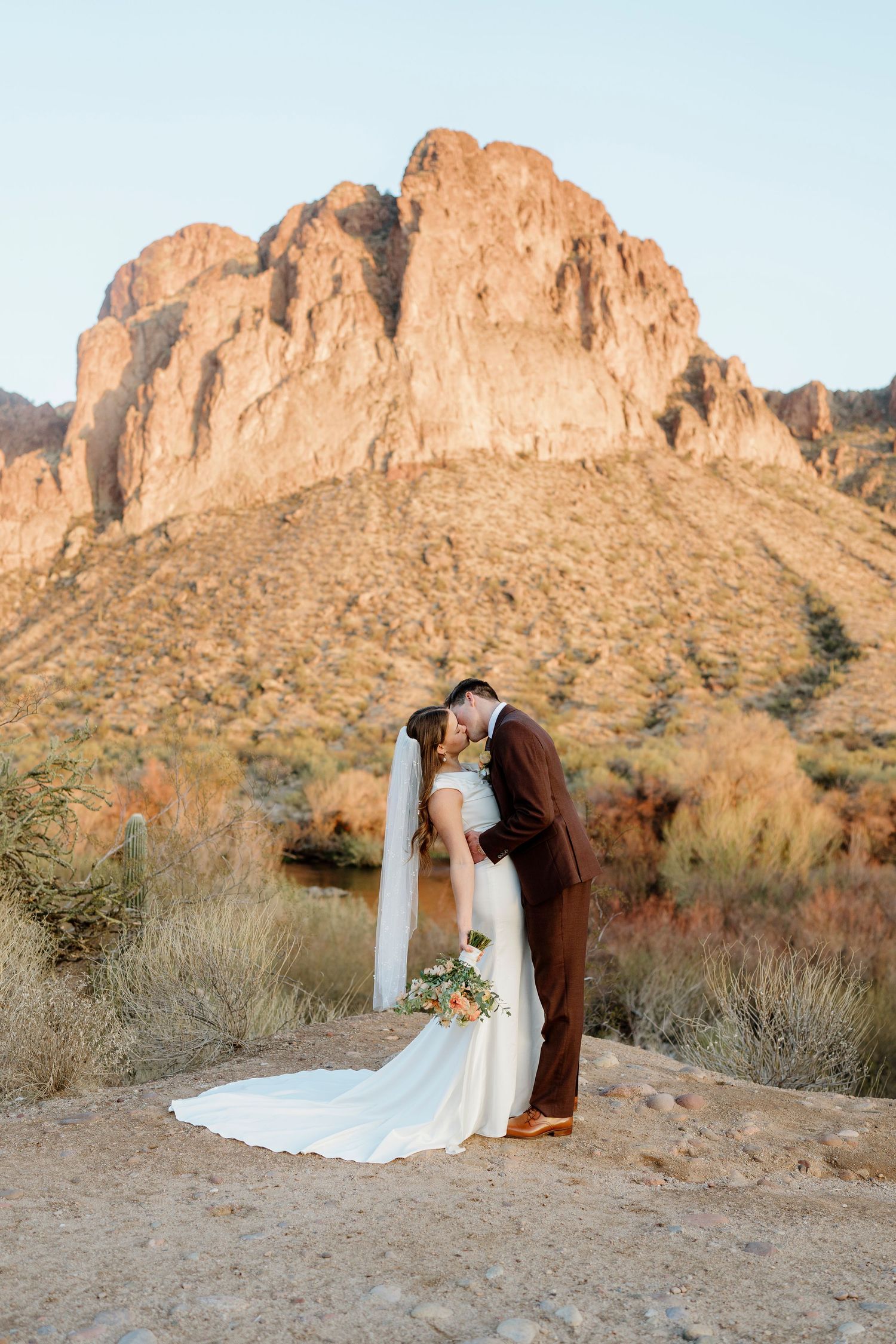 Bride and groom kissing with dramatic rocky mountain backdrop at warm desert sunset.