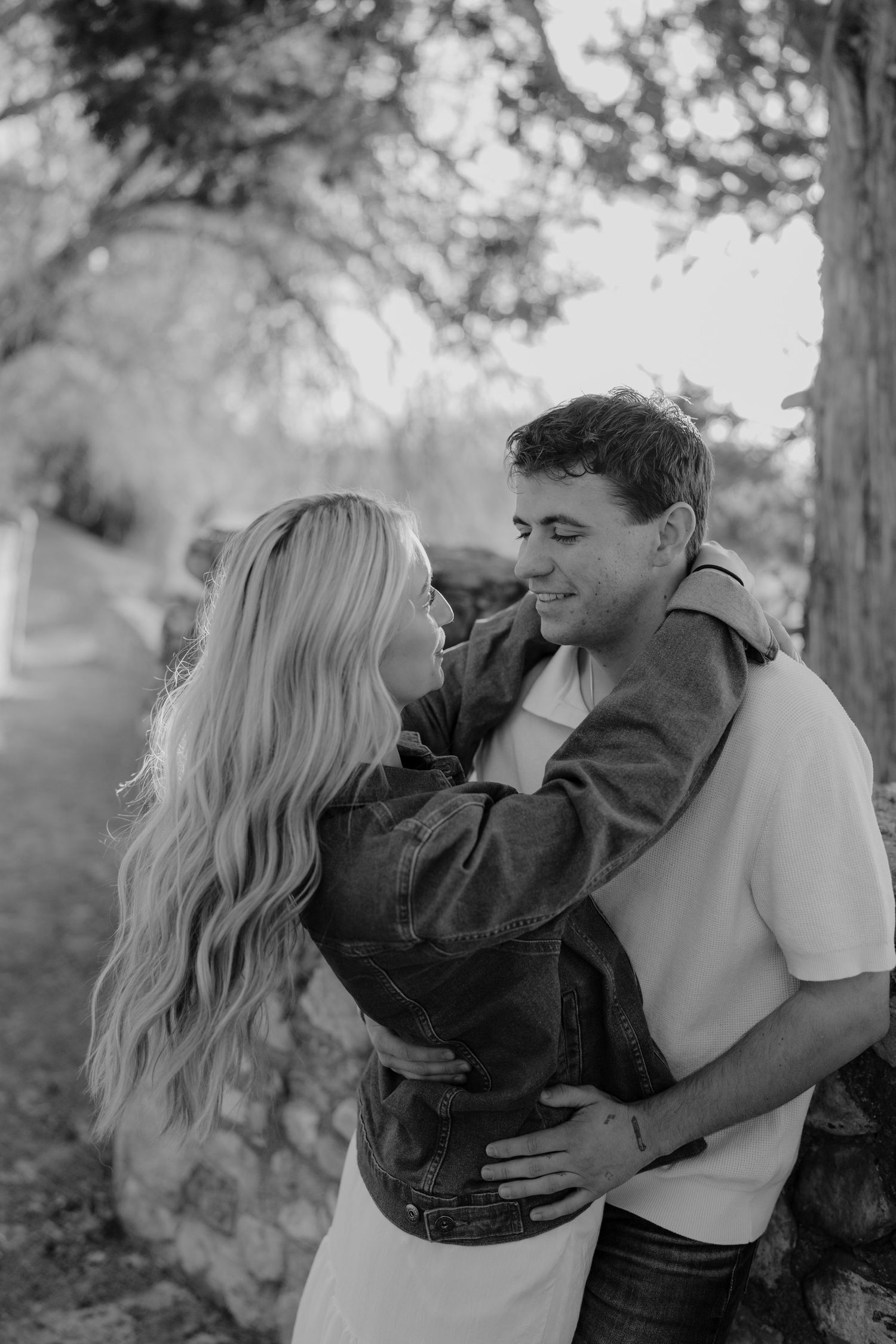 Black and white photo of a couple embracing tenderly under a large tree in an outdoor setting.