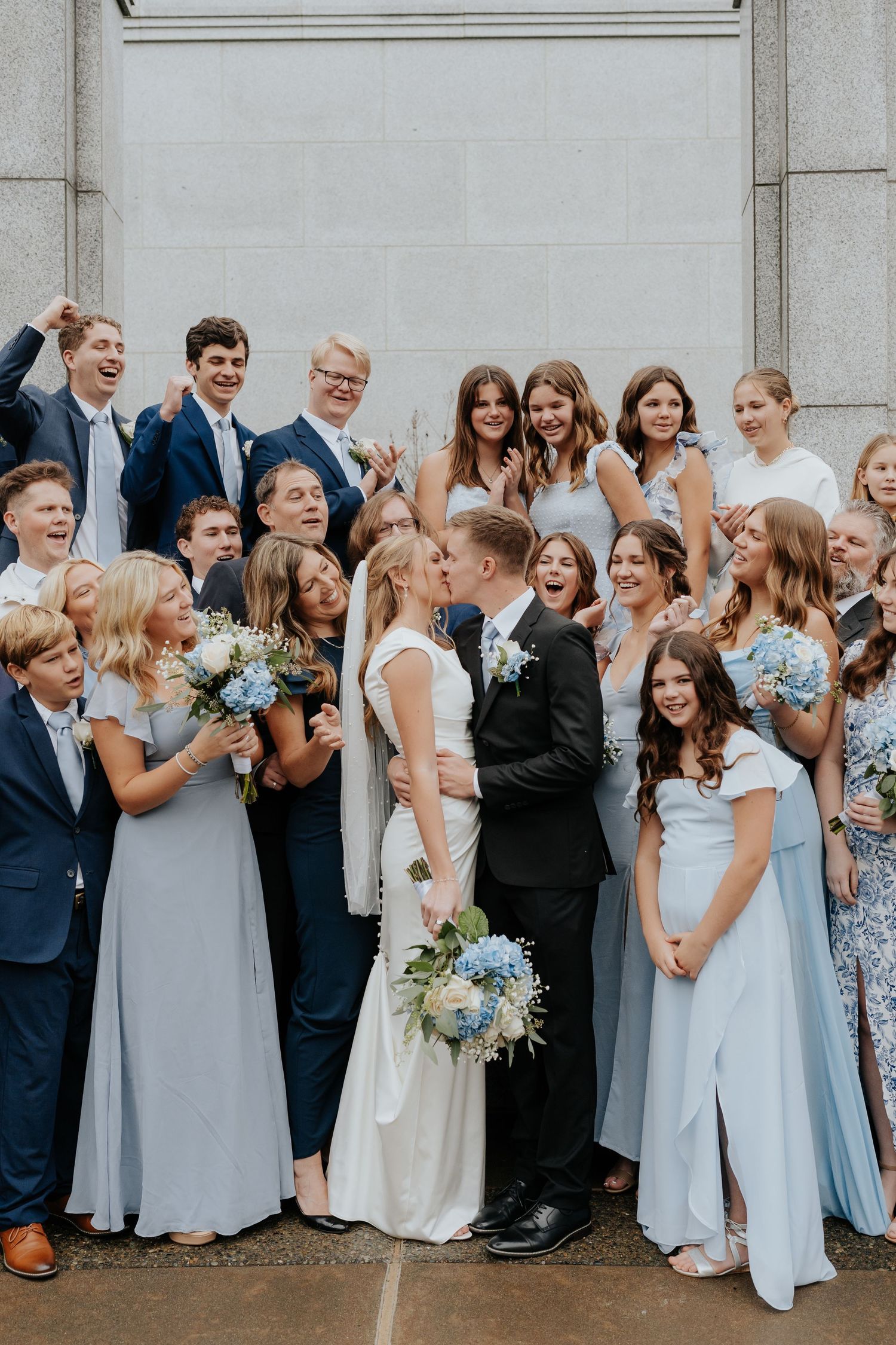 Large wedding party group celebrating with bride and groom kissing outside Sacramento LDS temple.