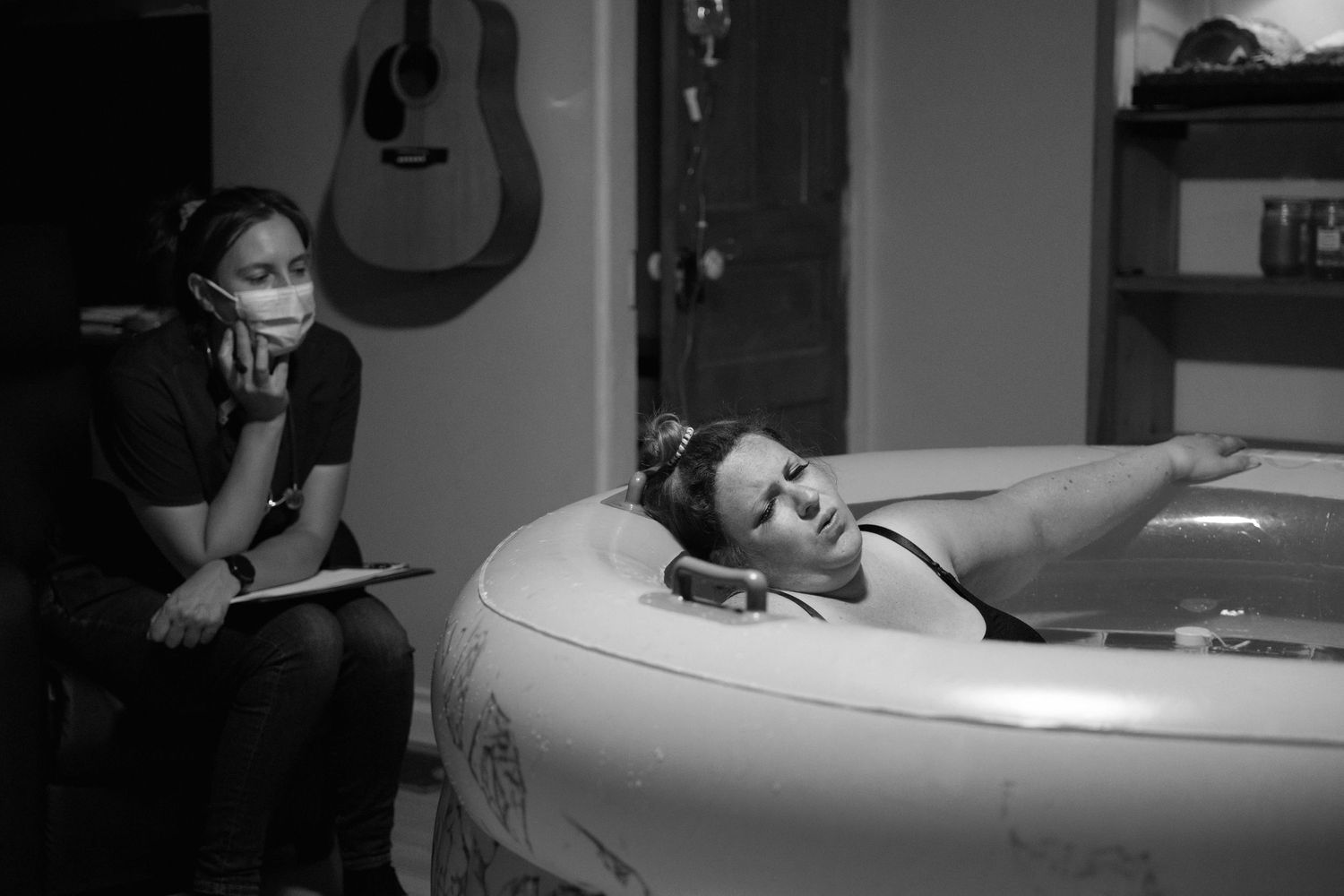 A black and white photo shows someone relaxing in an inflatable birthing pool while another person sits nearby.