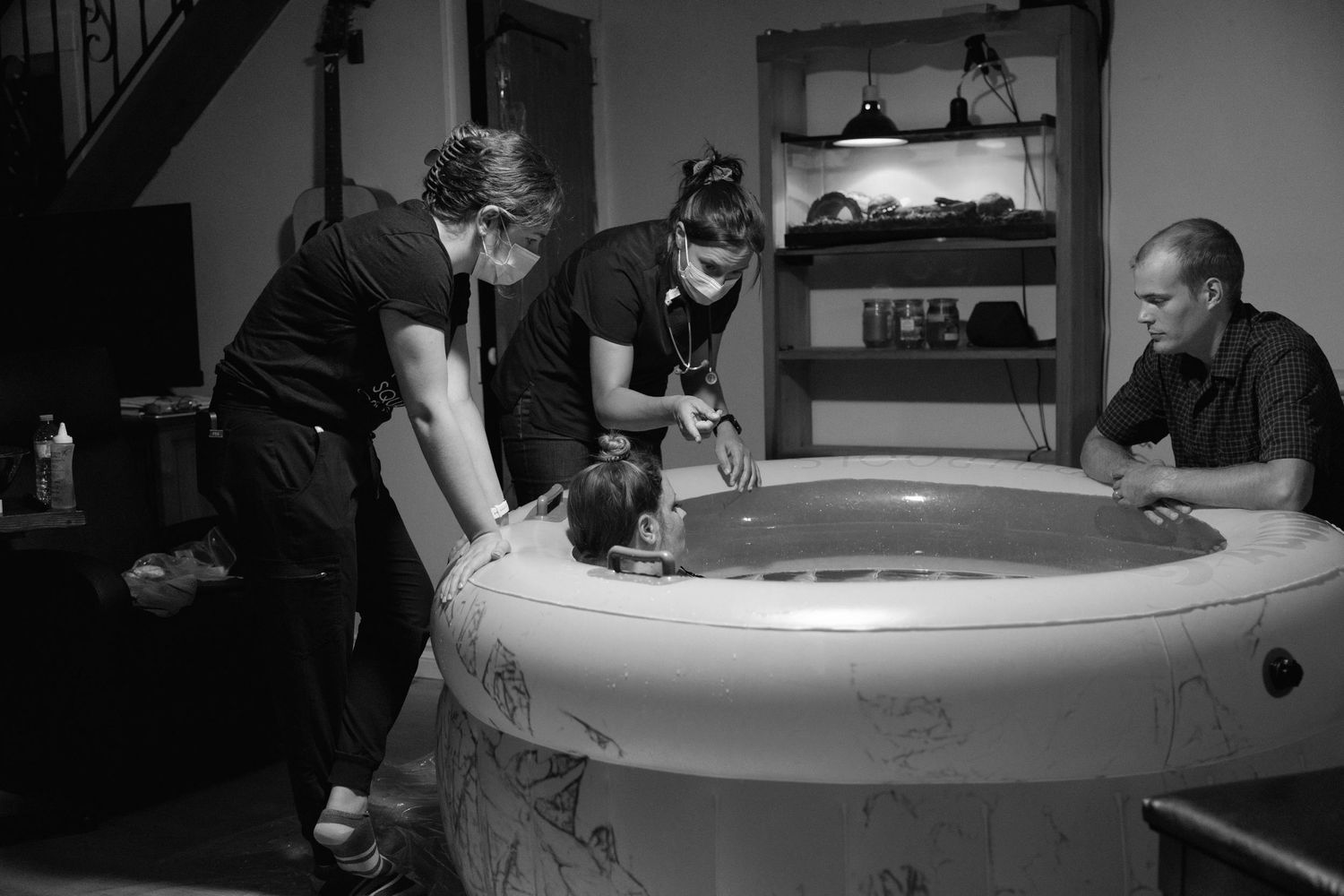 People gather around an inflatable birth pool during what appears to be a home birth preparation in black and white.